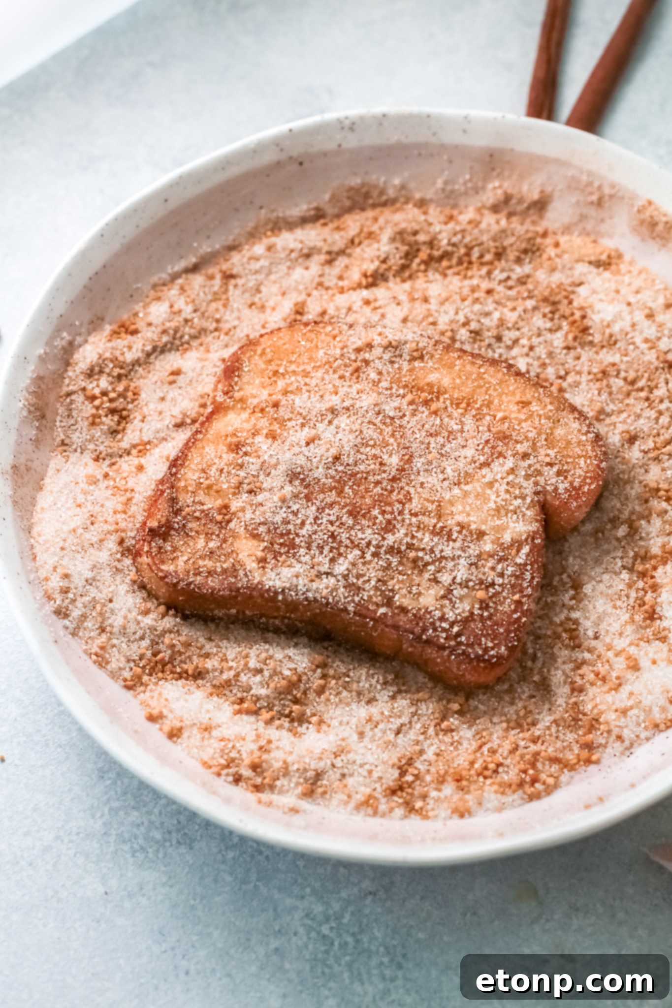 French toast being coated in a bowl of cinnamon sugar, showing a generous and even distribution.