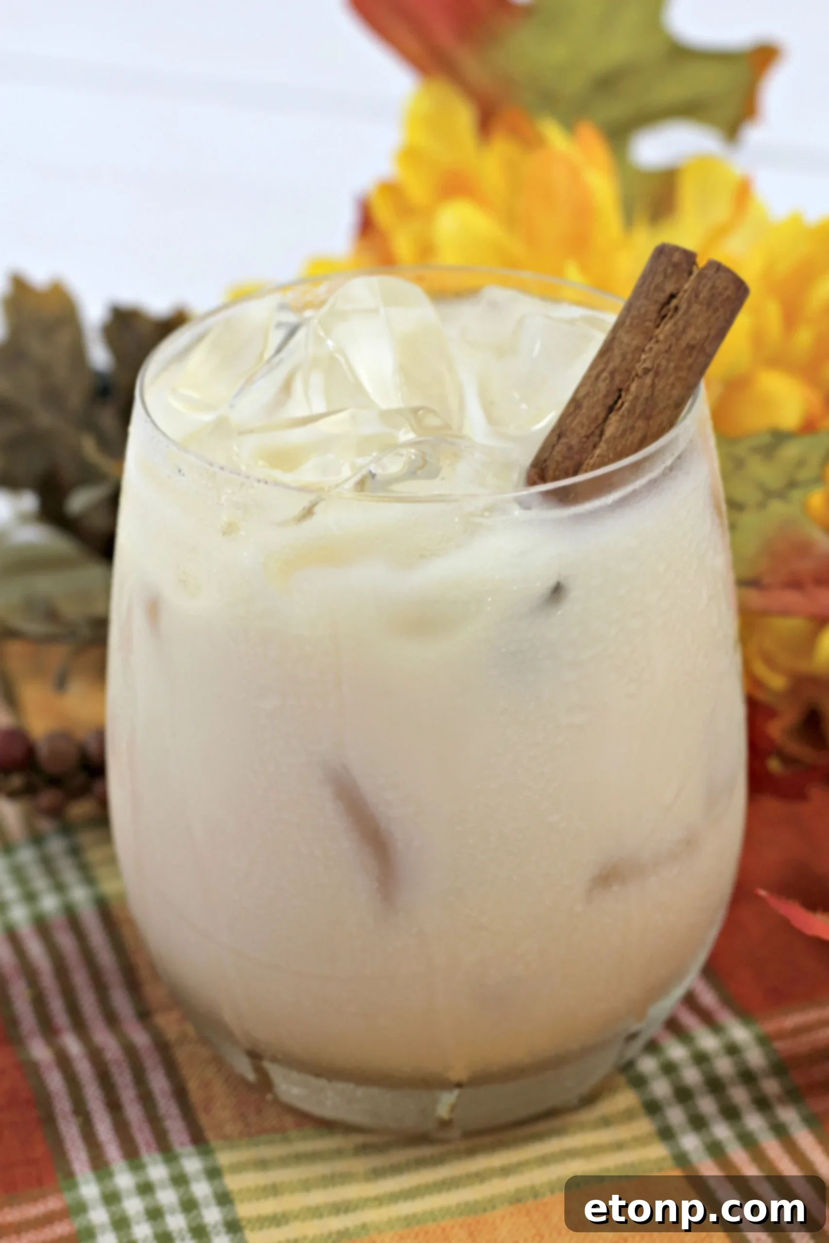 Overhead view of a hand holding a Pumpkin Spice White Russian, with a cozy blanket and fireplace in the background, creating a warm fall atmosphere.