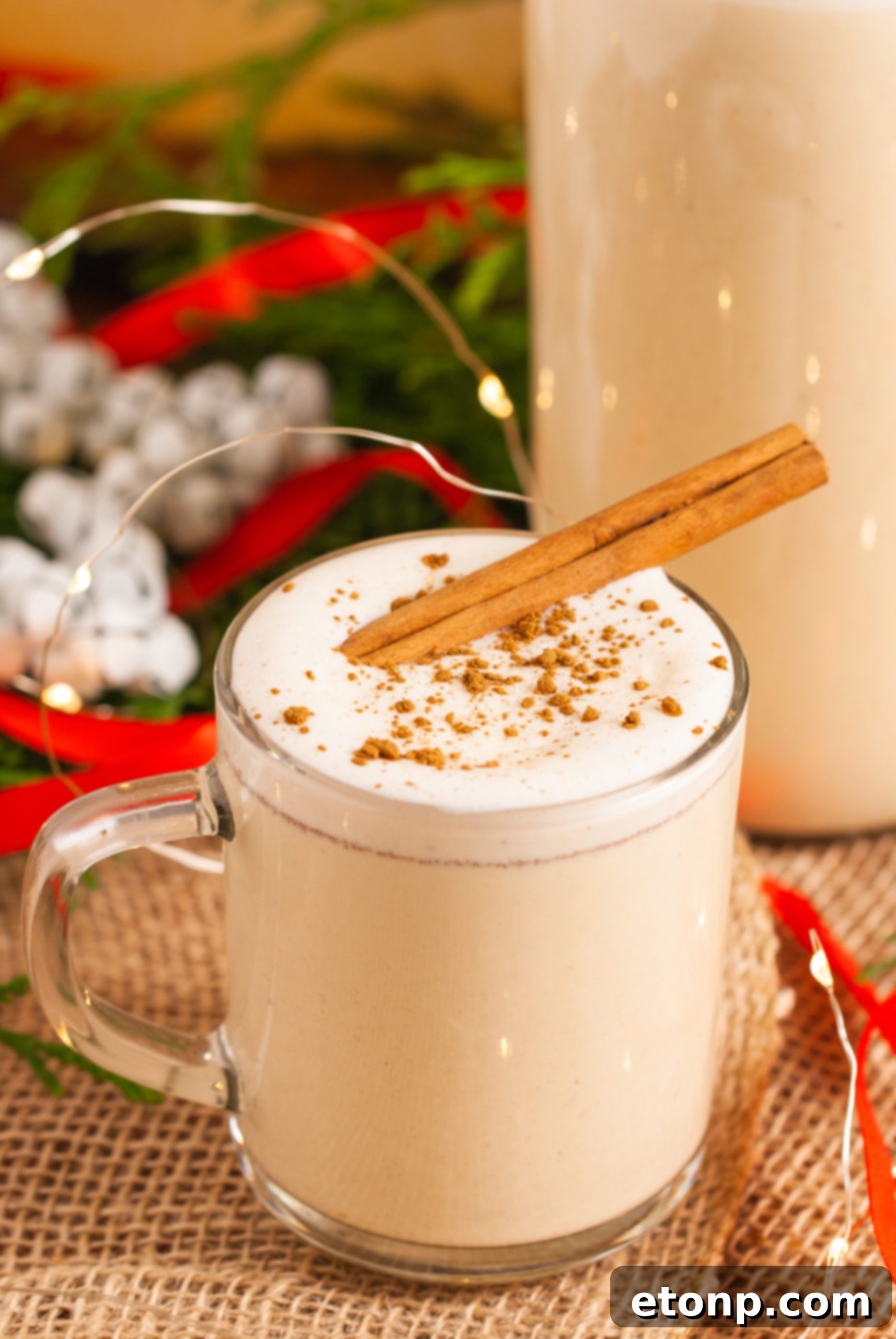 Two glasses of eggnog, one plain and one garnished, sitting on a wooden surface with holiday decorations.