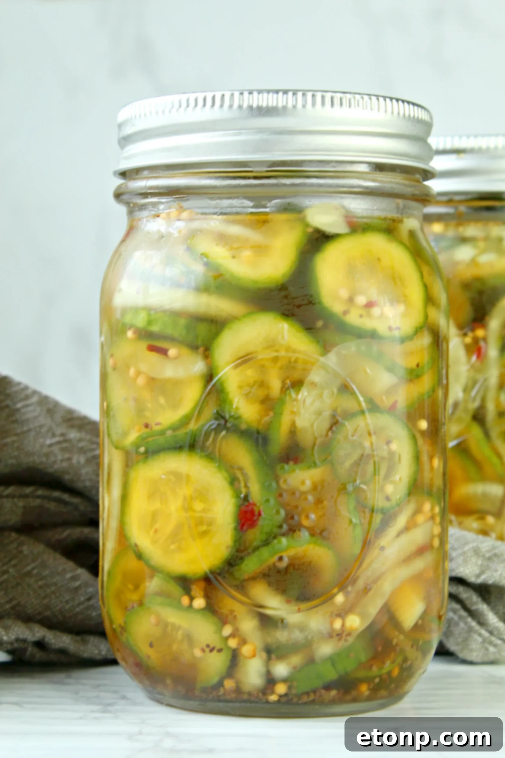 A close-up shot of freshly made refrigerator bread and butter pickles, thinly sliced and perfectly brined, overflowing from a glass jar with a rustic wooden background.