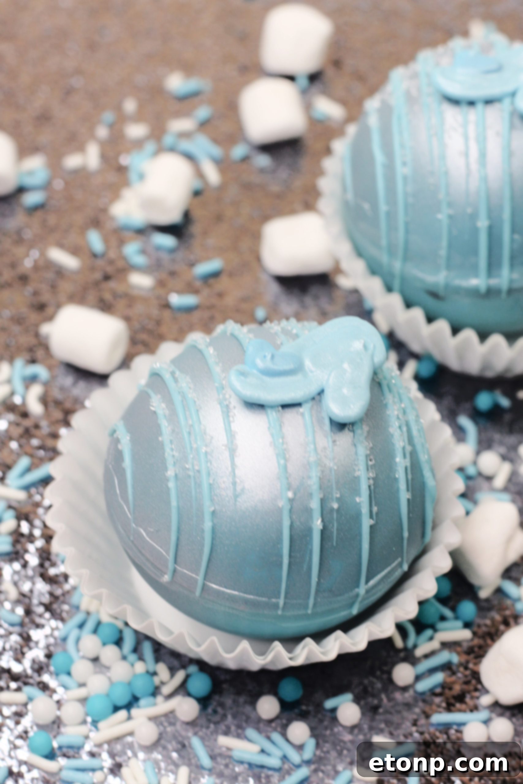 A close-up of a Cinderella Hot Cocoa Bomb, highlighting its smooth, glossy blue chocolate shell and the shimmering silver accents, alongside a small chocolate glass slipper.