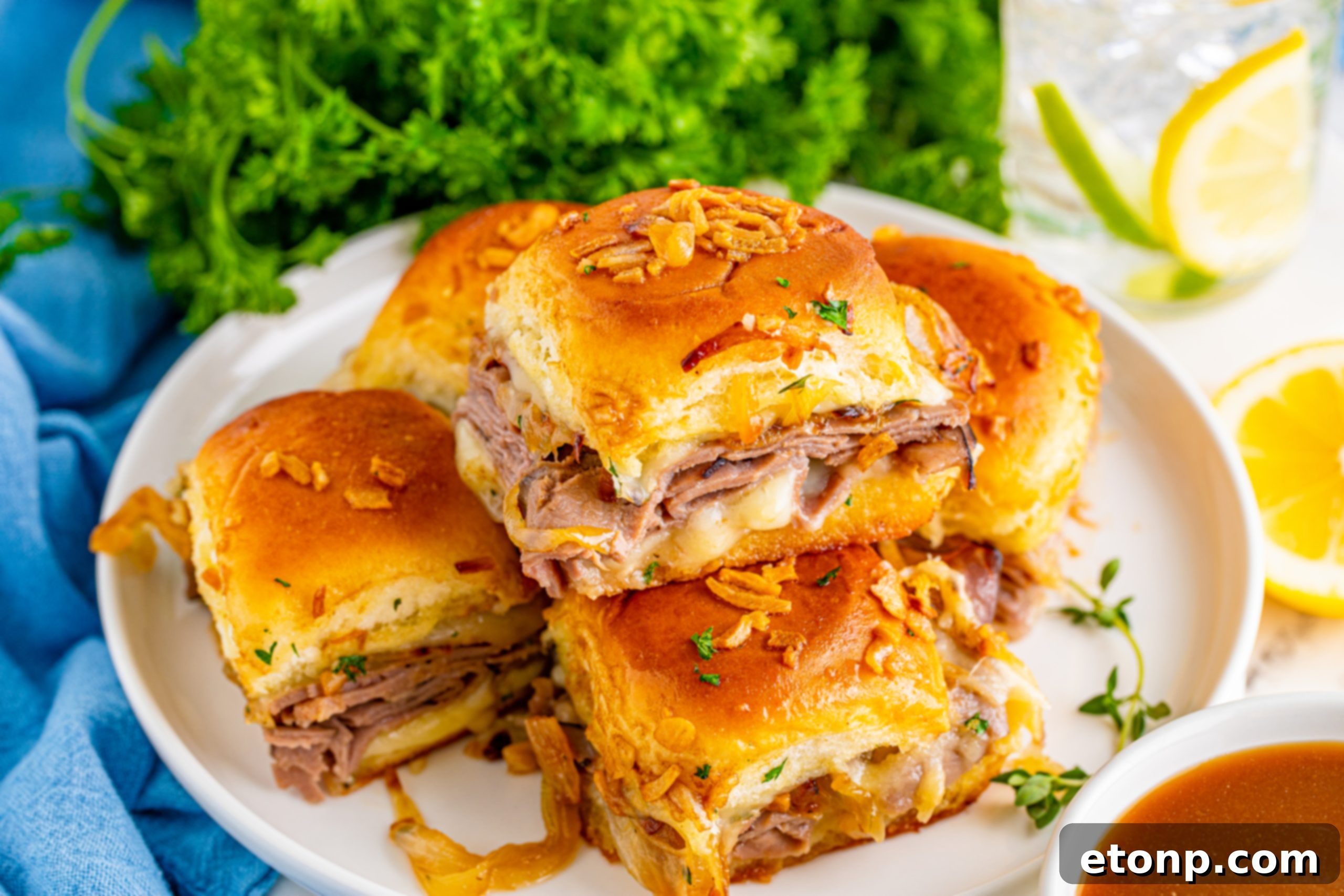 Several French Dip Sliders piled on a white plate next to a blue napkin.