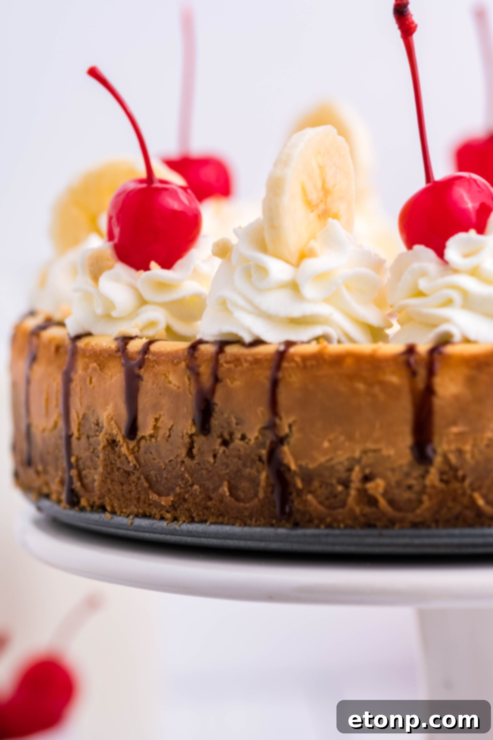 A full Banana Split Cheesecake, beautifully decorated with whipped cream, chocolate syrup, banana slices, and cherries, ready to be sliced and served at a gathering.