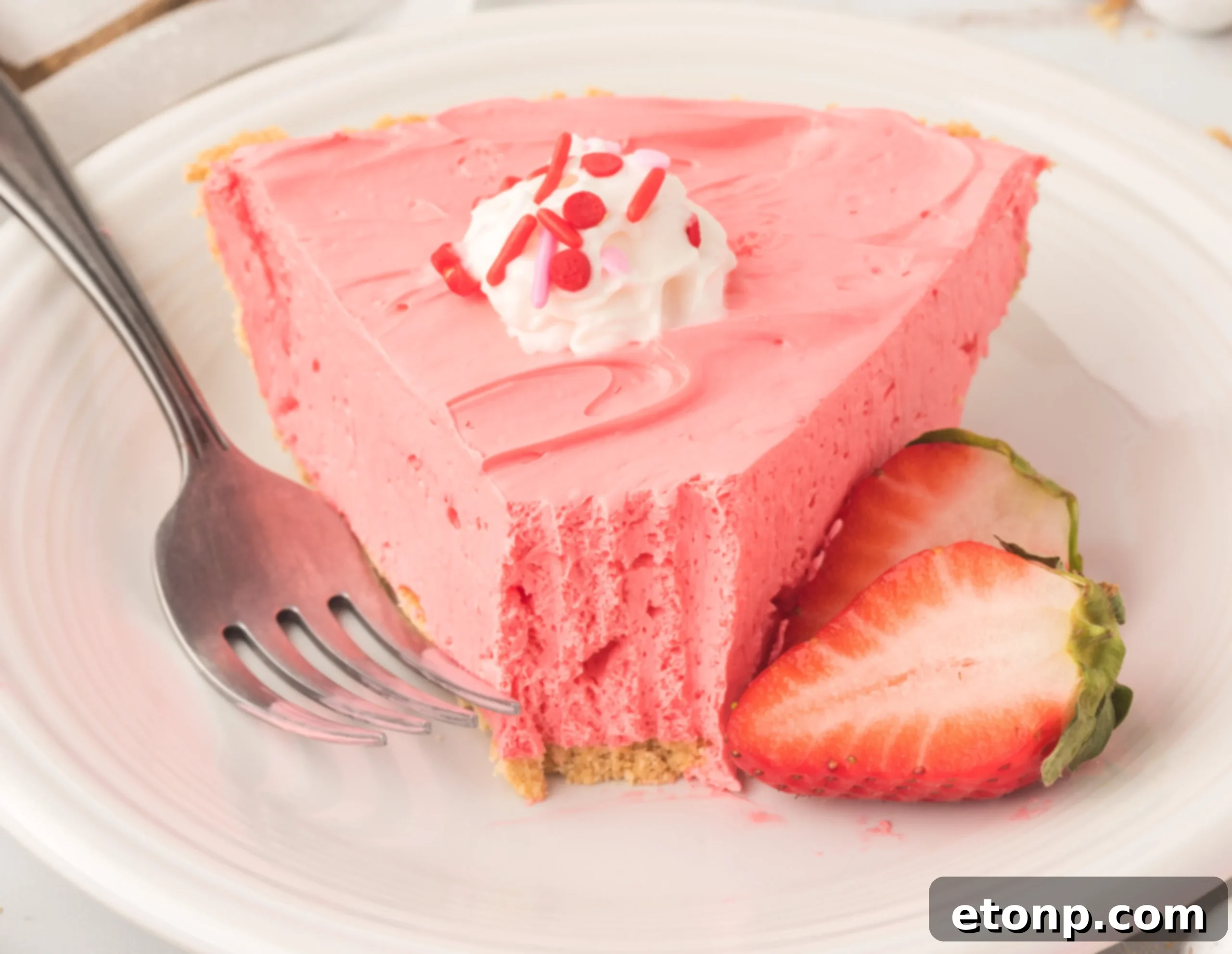 A vibrant pink slice of no-bake Strawberry Kool-Aid pie, garnished with fresh whipped cream and ripe strawberries, served on a white plate.