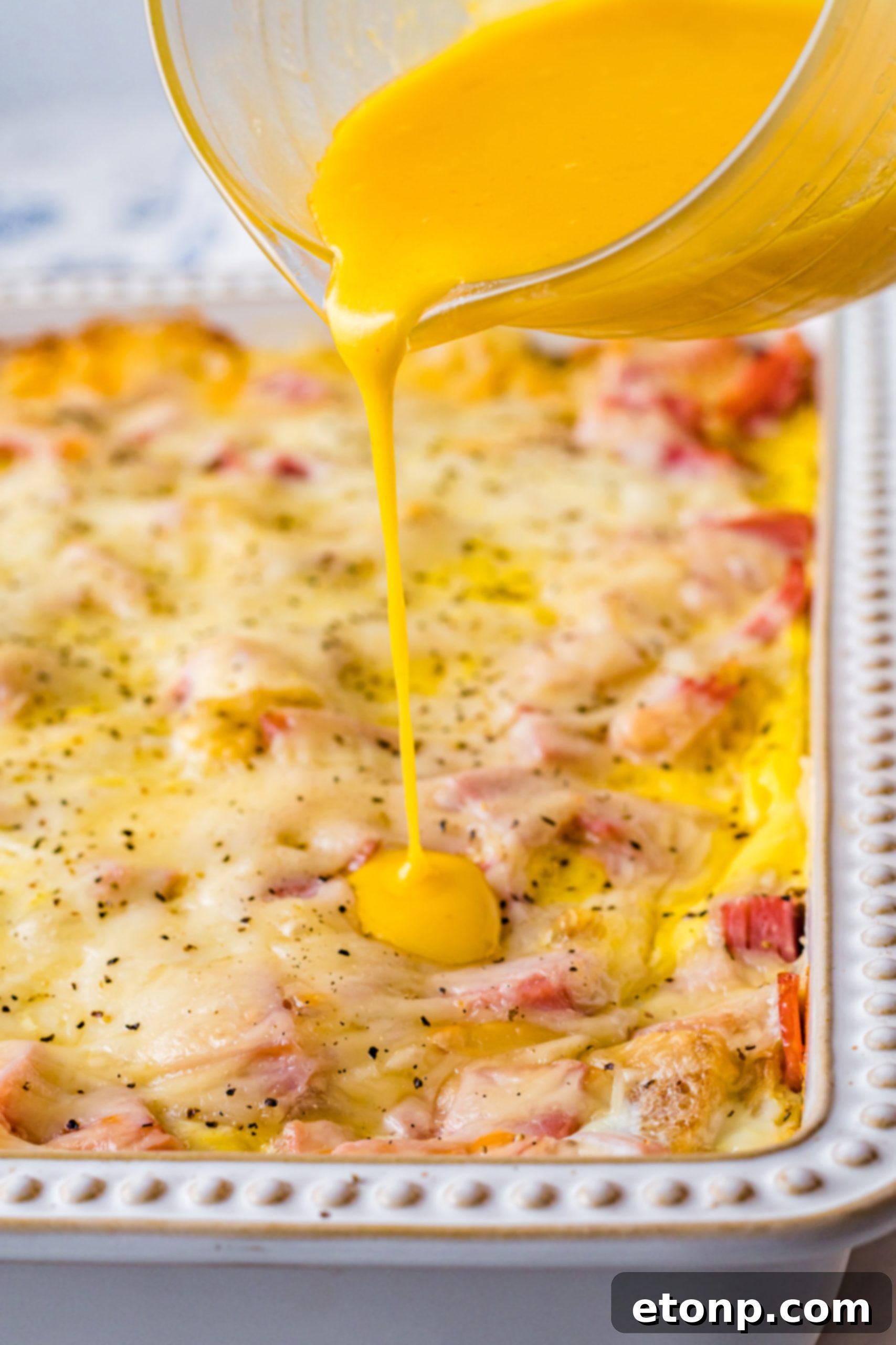 Eggs Benedict in a white casserole dish with hollandaise sauce being poured on top