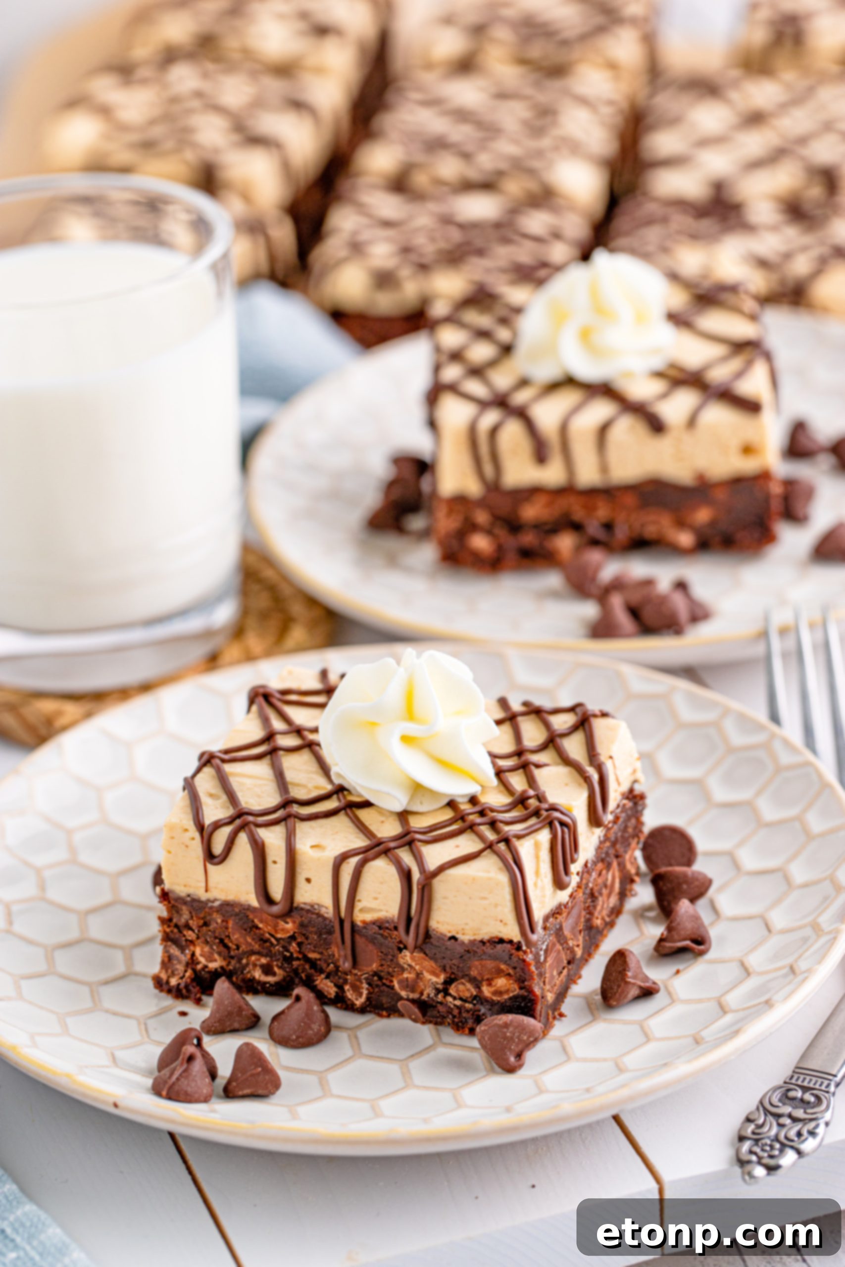 Peanut Butter Brownies on a white plate drizzled with chocolate and topped with whipped cream with a cup of milk beside it and chocolate morsels on the plate