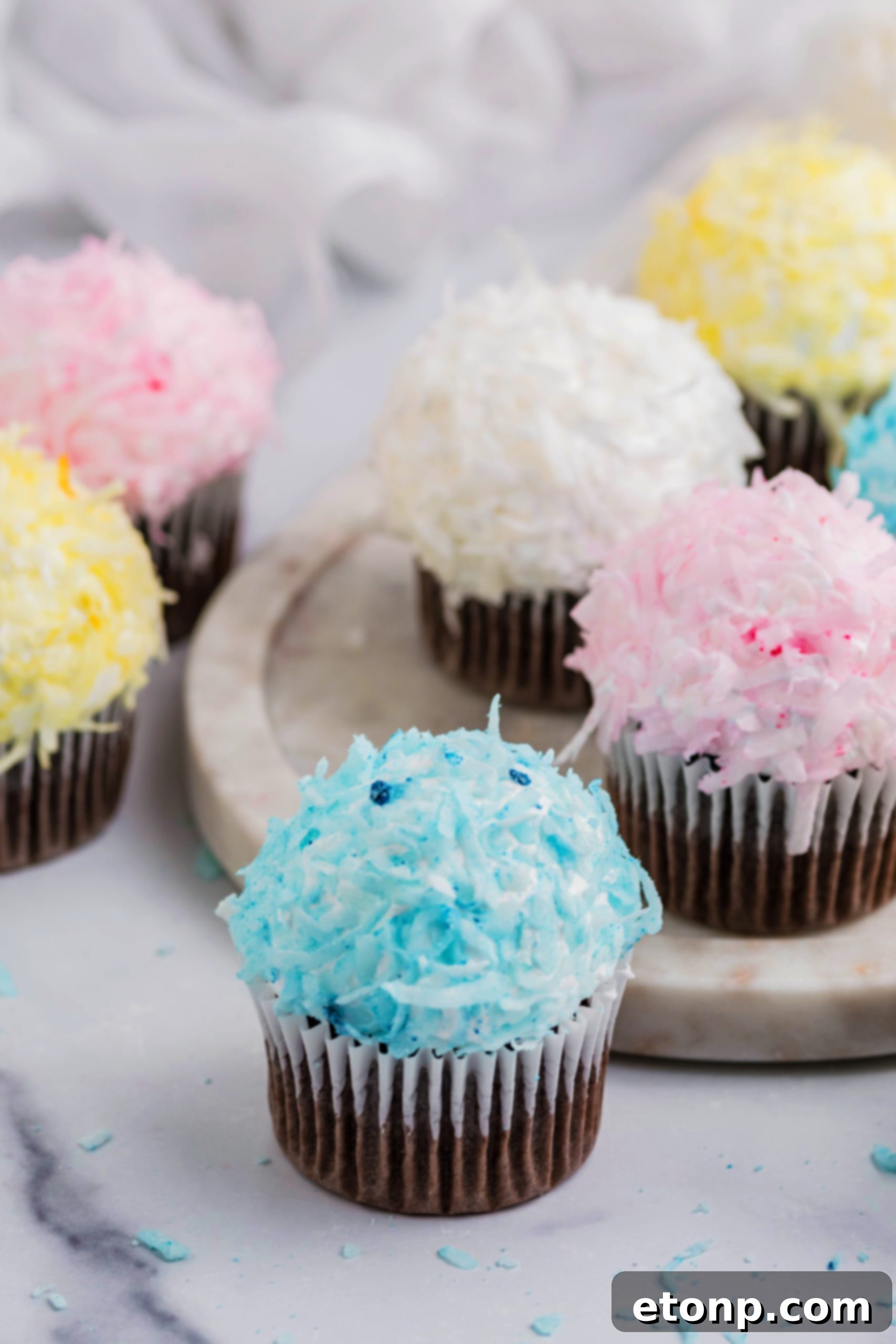 A vibrant display of pink, yellow, white, and blue chocolate cupcakes generously topped with fluffy marshmallow frosting and colorful coconut flakes, artfully arranged to resemble adorable bunny butts.