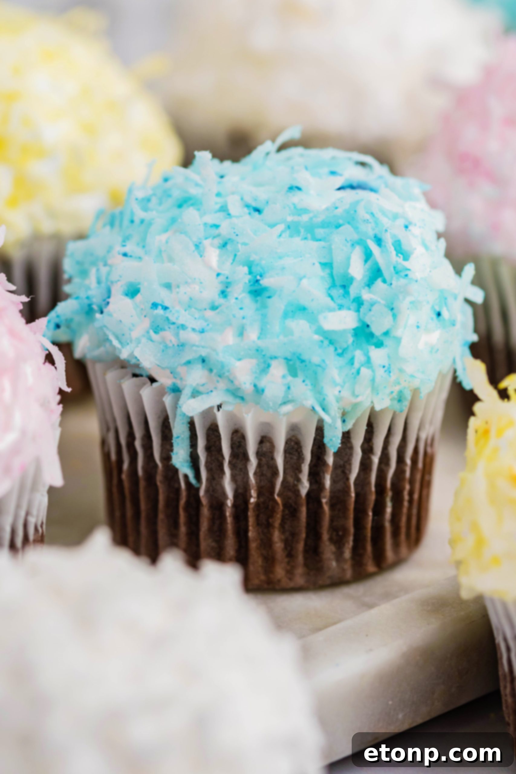 A colorful assortment of chocolate cupcakes, each crowned with vibrant pink, yellow, white, and blue coconut flakes and marshmallow frosting, creating a festive Easter treat.