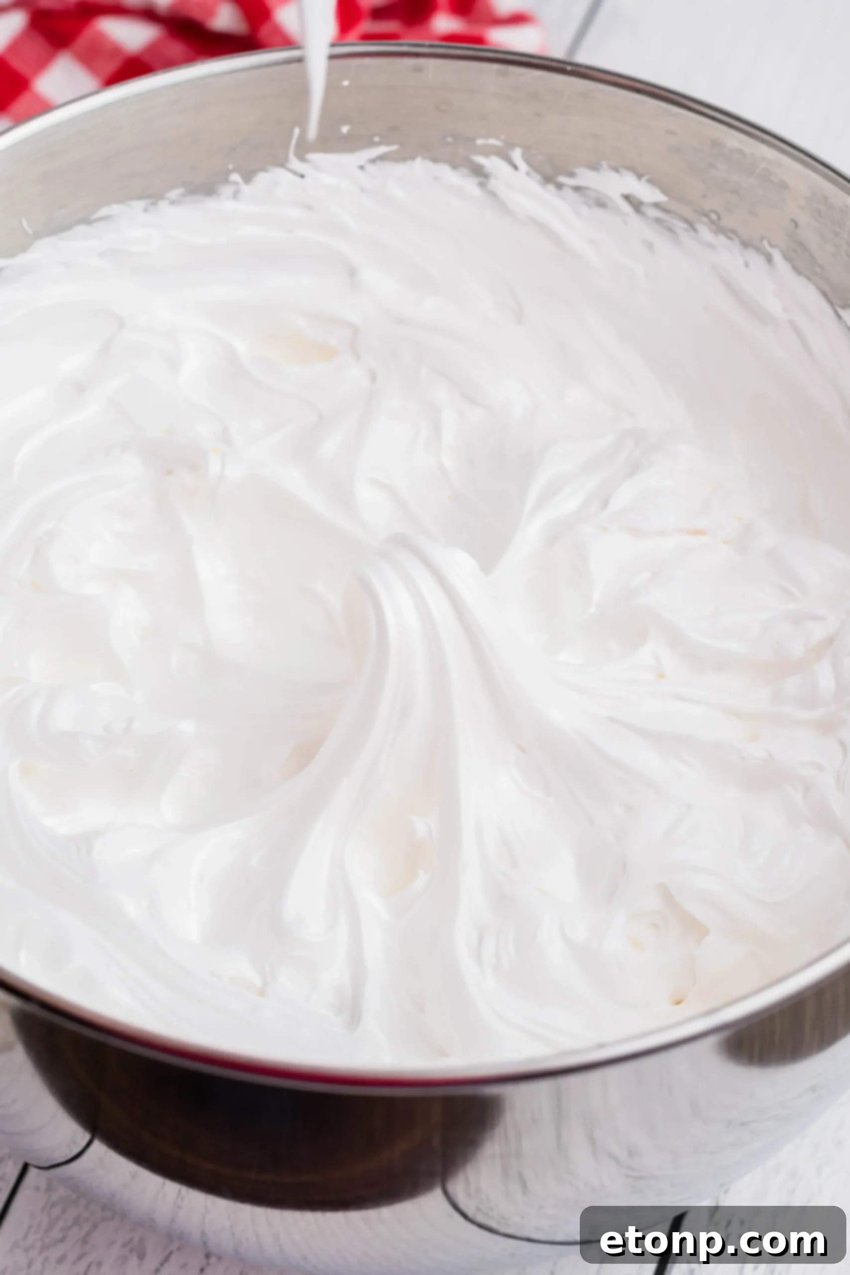 A close-up shot of homemade marshmallow frosting, showcasing its silky smooth and glossy texture, in a gleaming stainless steel mixing bowl.