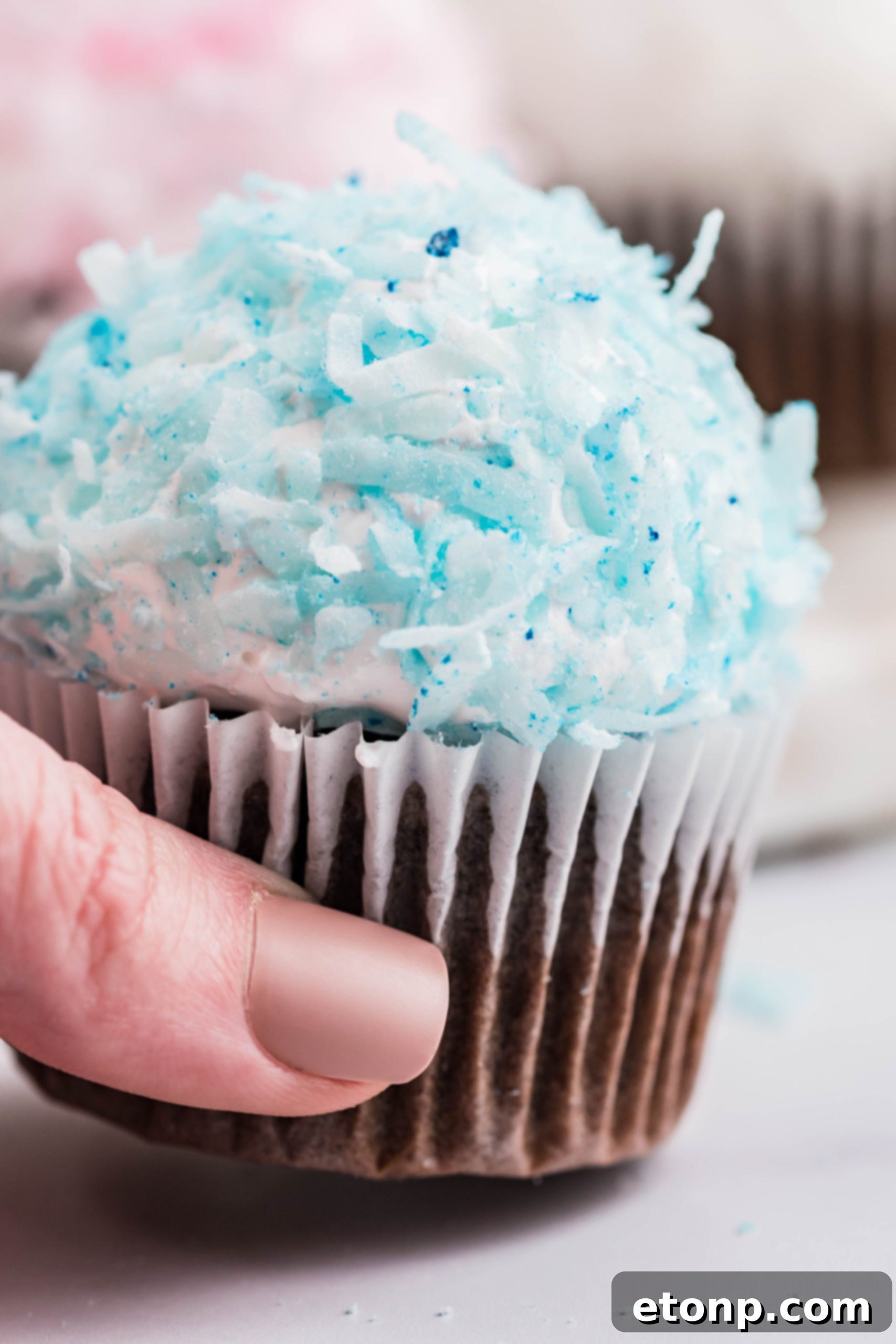 A hand gently lifts a perfectly crafted chocolate cupcake, showcasing its vibrant blue coconut shavings and cloud-like marshmallow frosting against a soft, inviting backdrop.