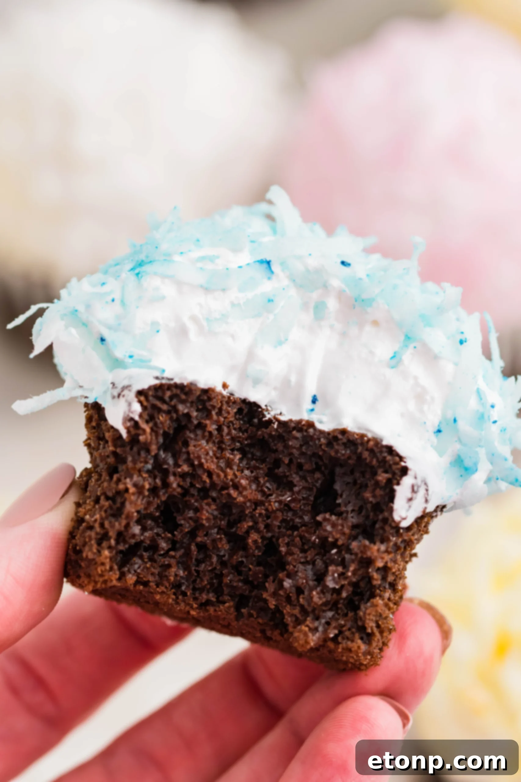 A chocolate cupcake with fluffy blue coconut shavings and marshmallow frosting, held up for a closer look, revealing a delectable bite taken out of it.