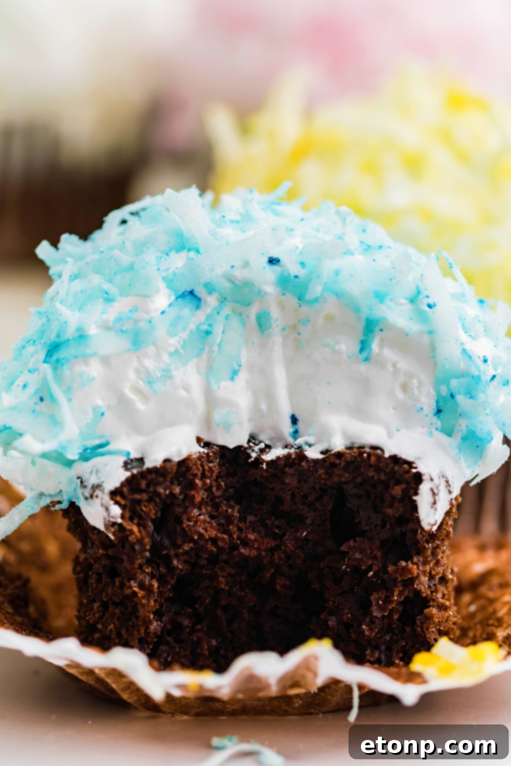 A close-up of a chocolate cupcake featuring delightful blue coconut shavings and marshmallow frosting, with a tempting bite already savored, highlighting its irresistible appeal.