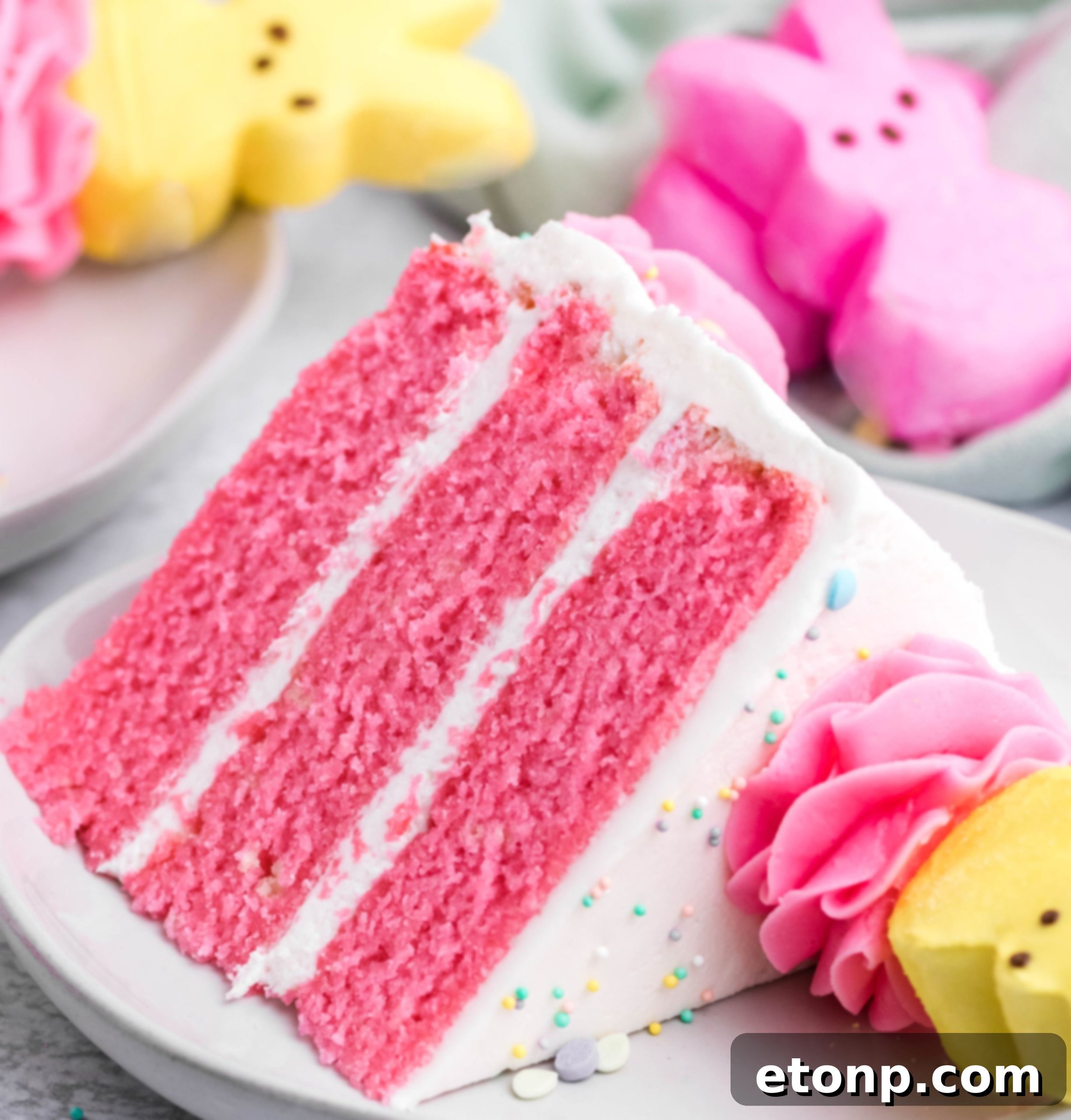 Pink Vanilla layered cake with white frosting and topped with yellow Peeps for Easter on a white plate with a slice removed