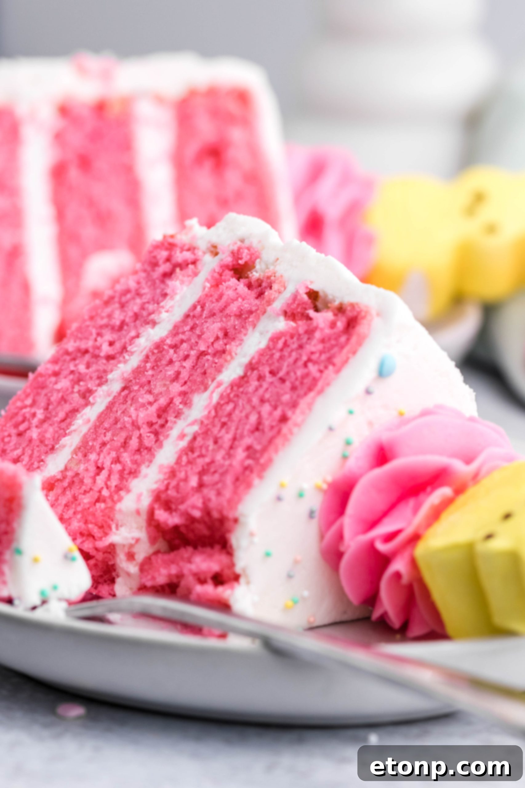 Pink Vanilla layered cake with white frosting and topped with yellow Peeps for Easter on a white plate with a slice gone