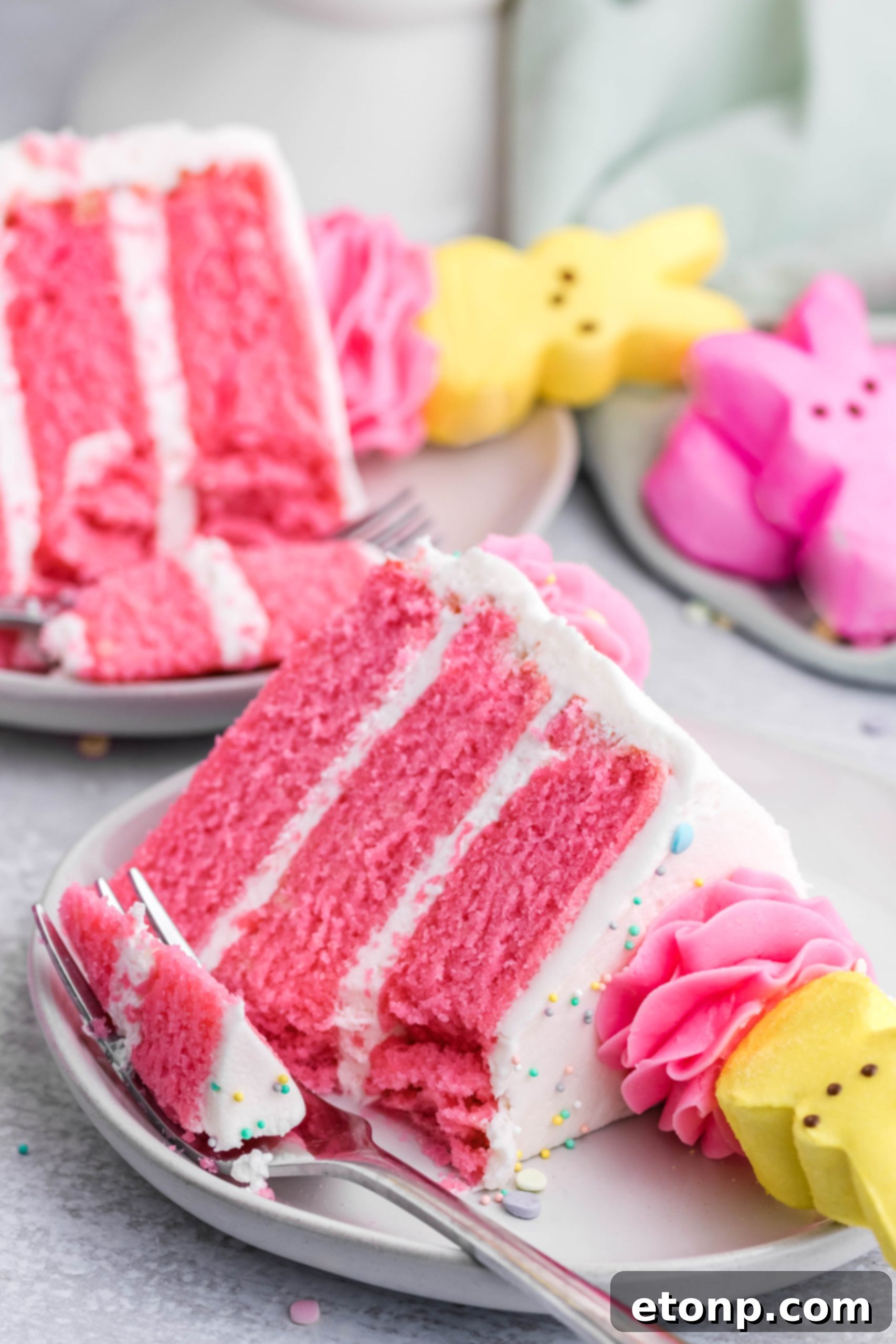 Pink Vanilla layered cake with white frosting and topped with yellow Peeps for Easter on a white plate with a slice gone