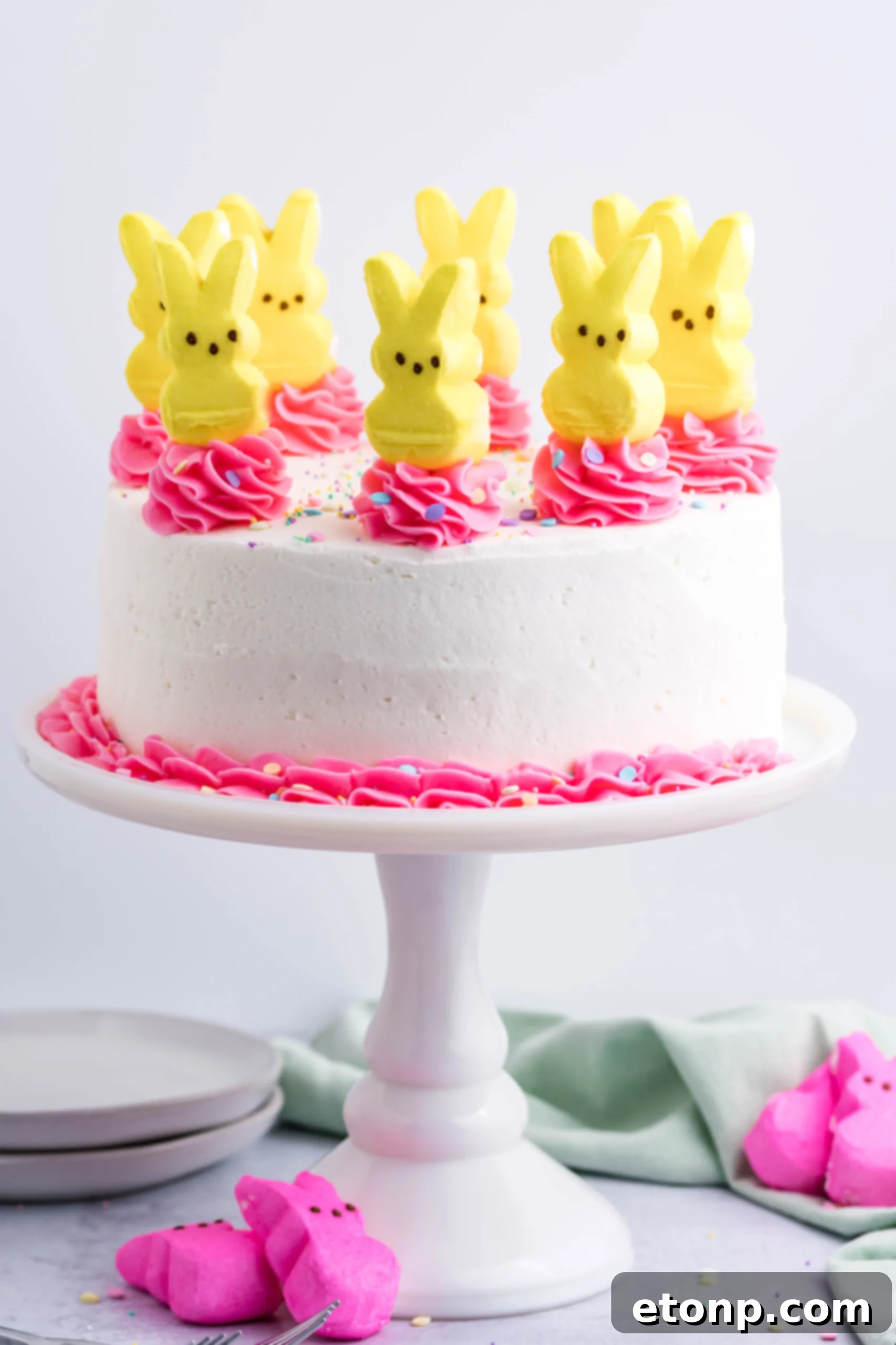 Pink Vanilla layered cake with white frosting and topped with yellow Peeps for Easter on a white cake stand