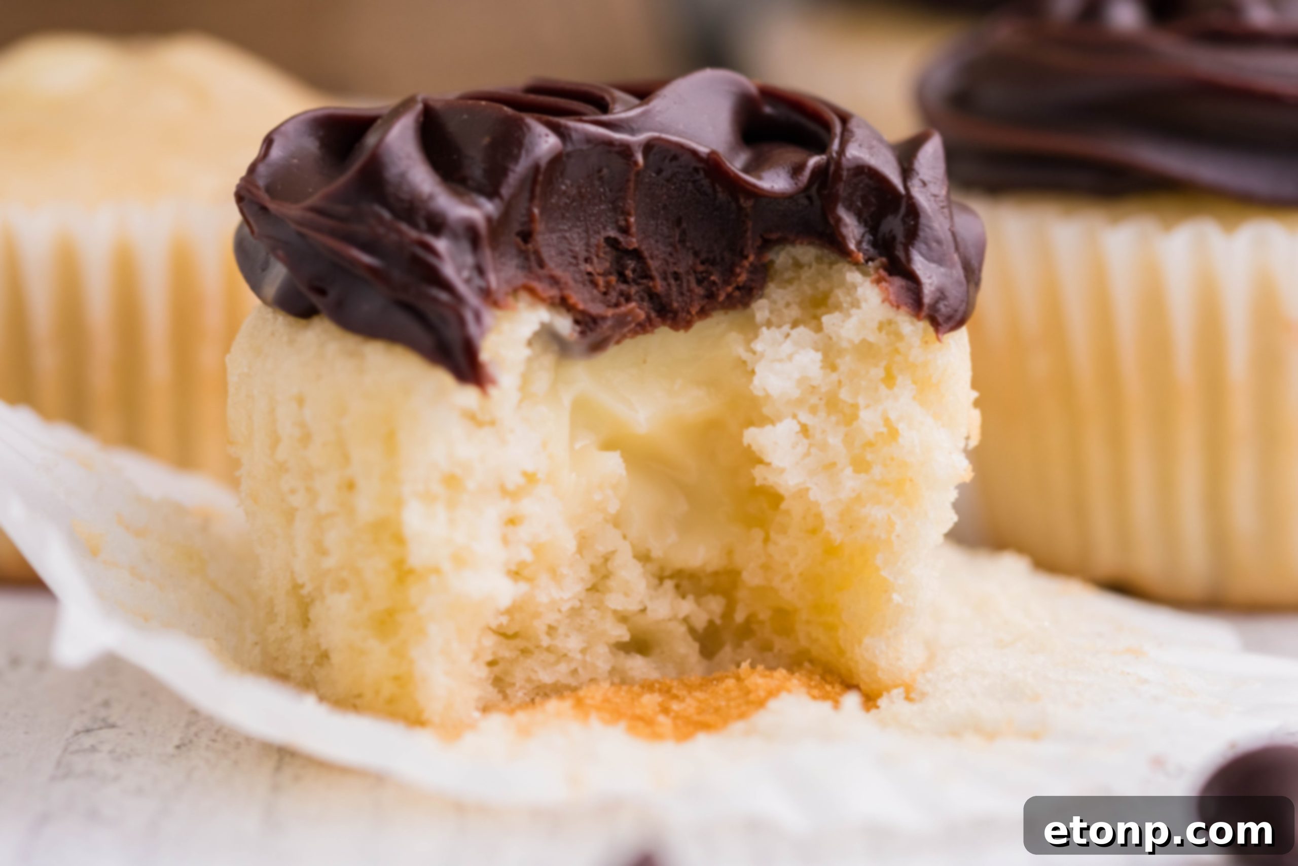 A Boston Cream Cupcake with a delightful bite missing, showcasing its layered perfection of cake, cream, and ganache on a white counter.