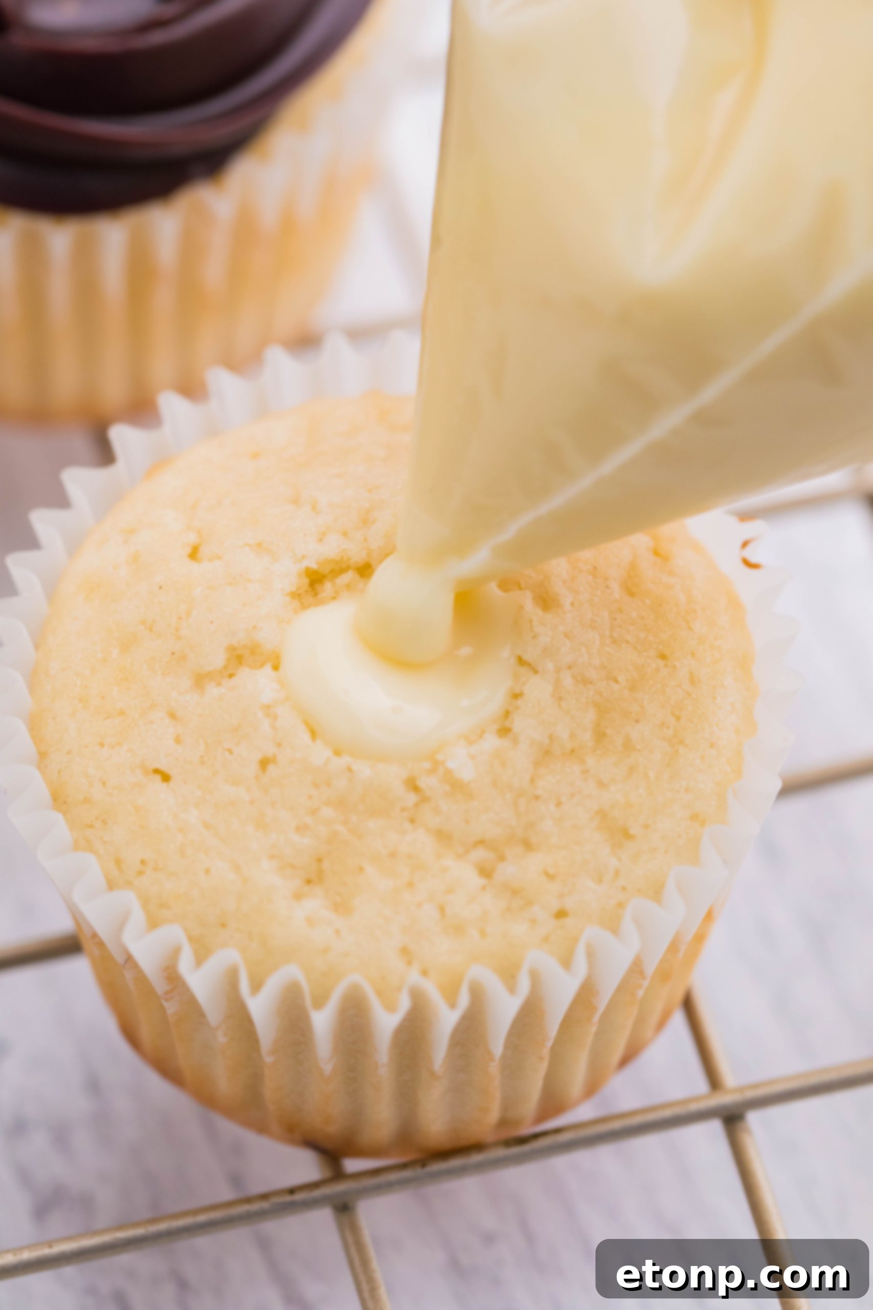 A piping bag precisely fills the hollowed center of a Boston Cream Cupcake with smooth, golden pastry cream.