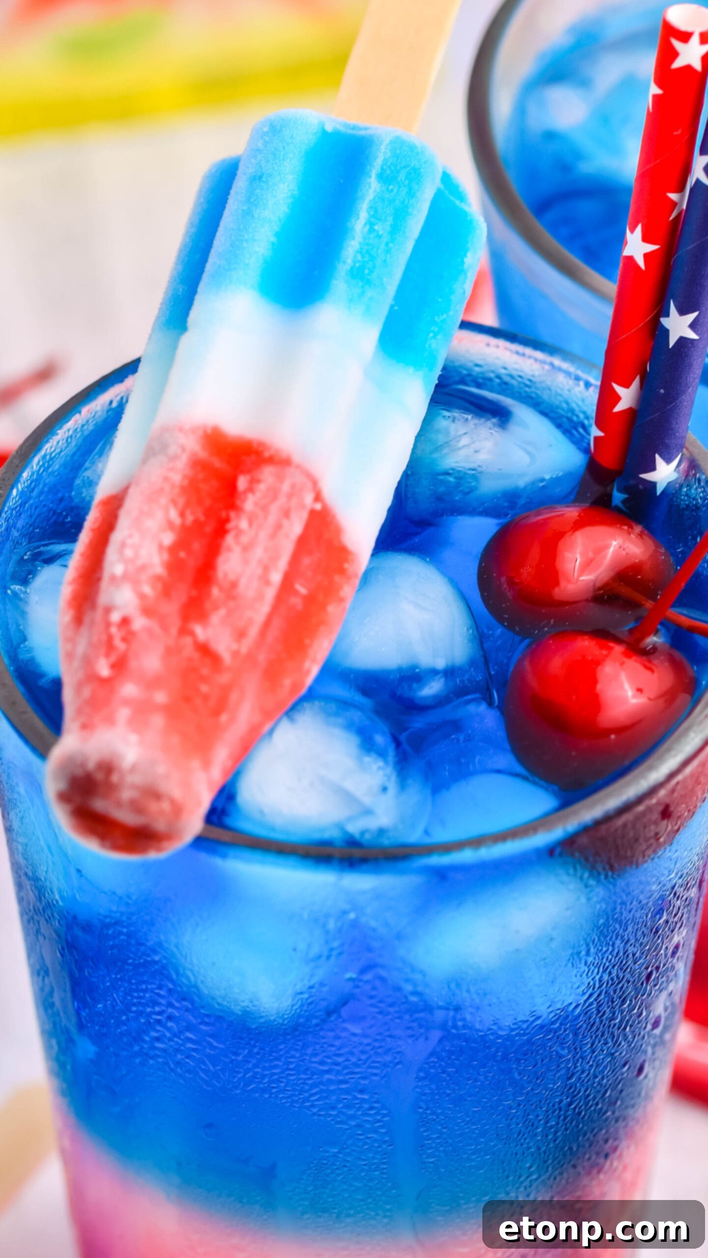 Bomb Pop Cocktail Drink in a clear glass with a blue & red straw with cherries floating up close