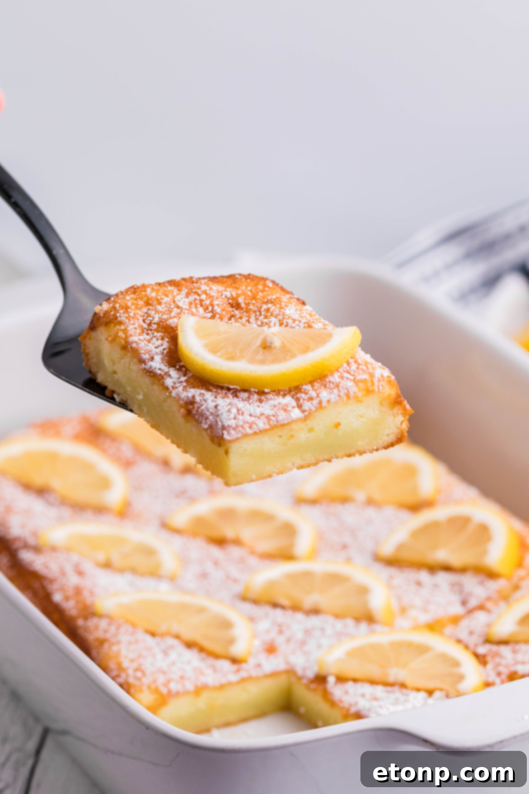 A slice of 2-Ingredient Lemon Bar being carefully lifted from the white baking pan with a spatula, showcasing its moist texture