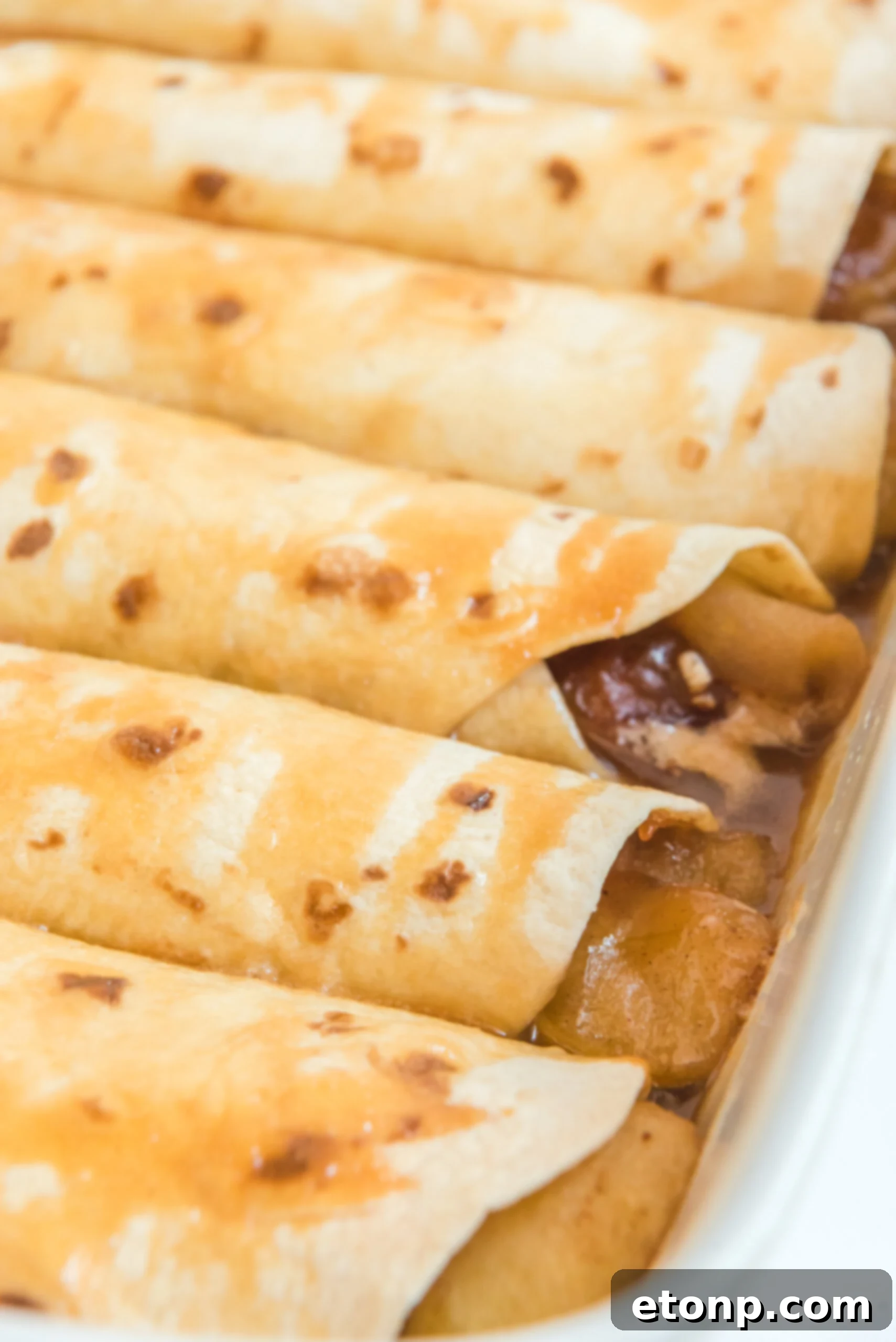 Cinnamon Apple Enchilada Bake 8 Apple Pie Enchiladas topped with caramel sauce in a white baking dish, side view
