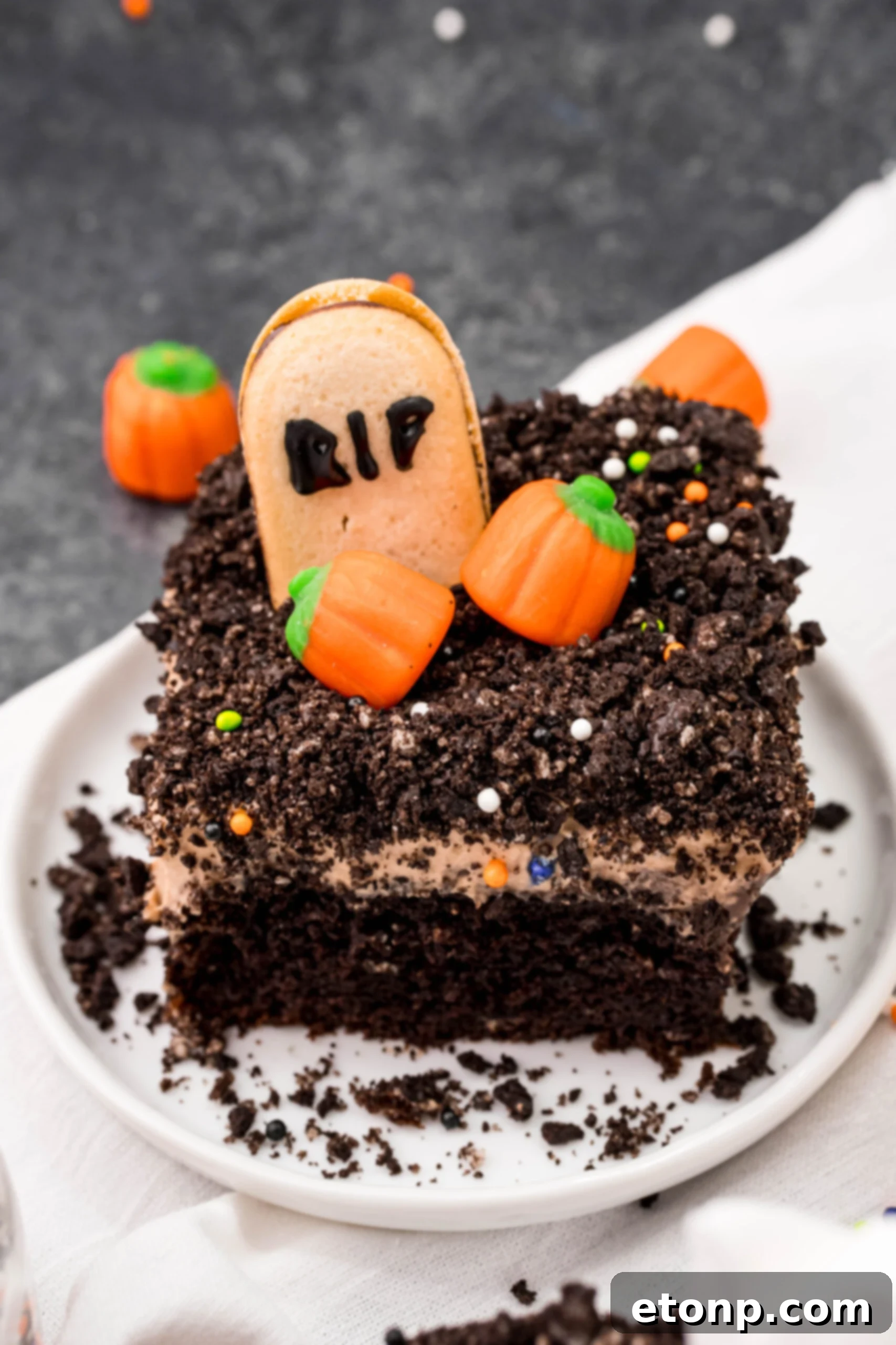 A slice of Halloween Dirt Cake on a white plate, garnished with Oreo crumbles and mini candy pumpkins, ready to be enjoyed.