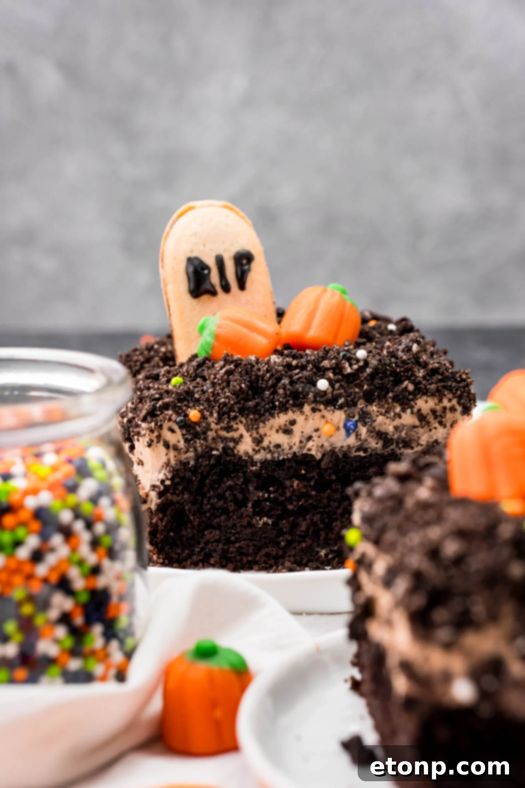 A slice of Halloween Dirt Cake on a white plate, accompanied by a glass jar filled with Halloween-colored sprinkles, adding a festive touch.
