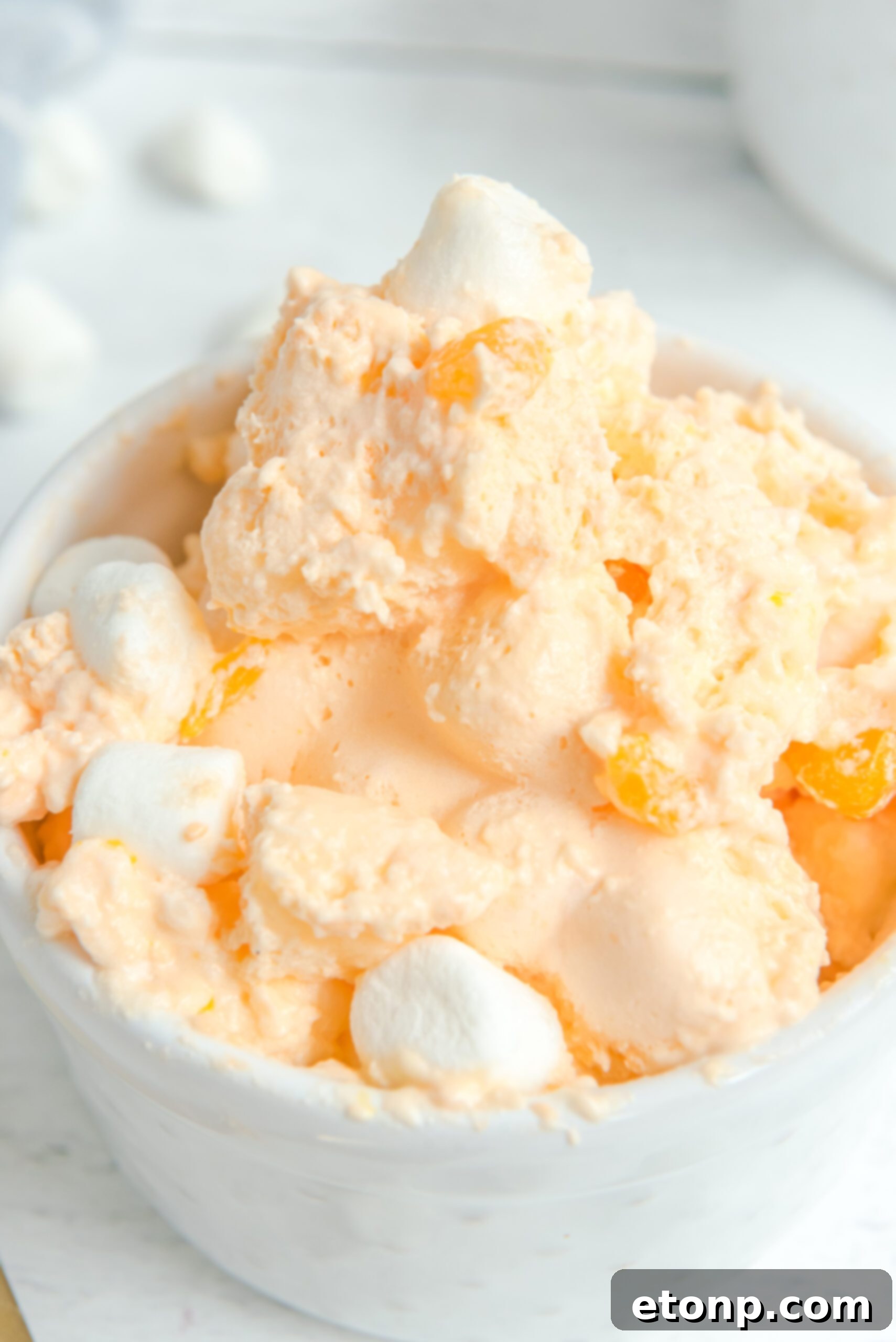 An extreme close-up of Orange Creamsicle Salad in a white bowl, artfully topped with mini marshmallows and vibrant mandarin orange segments.