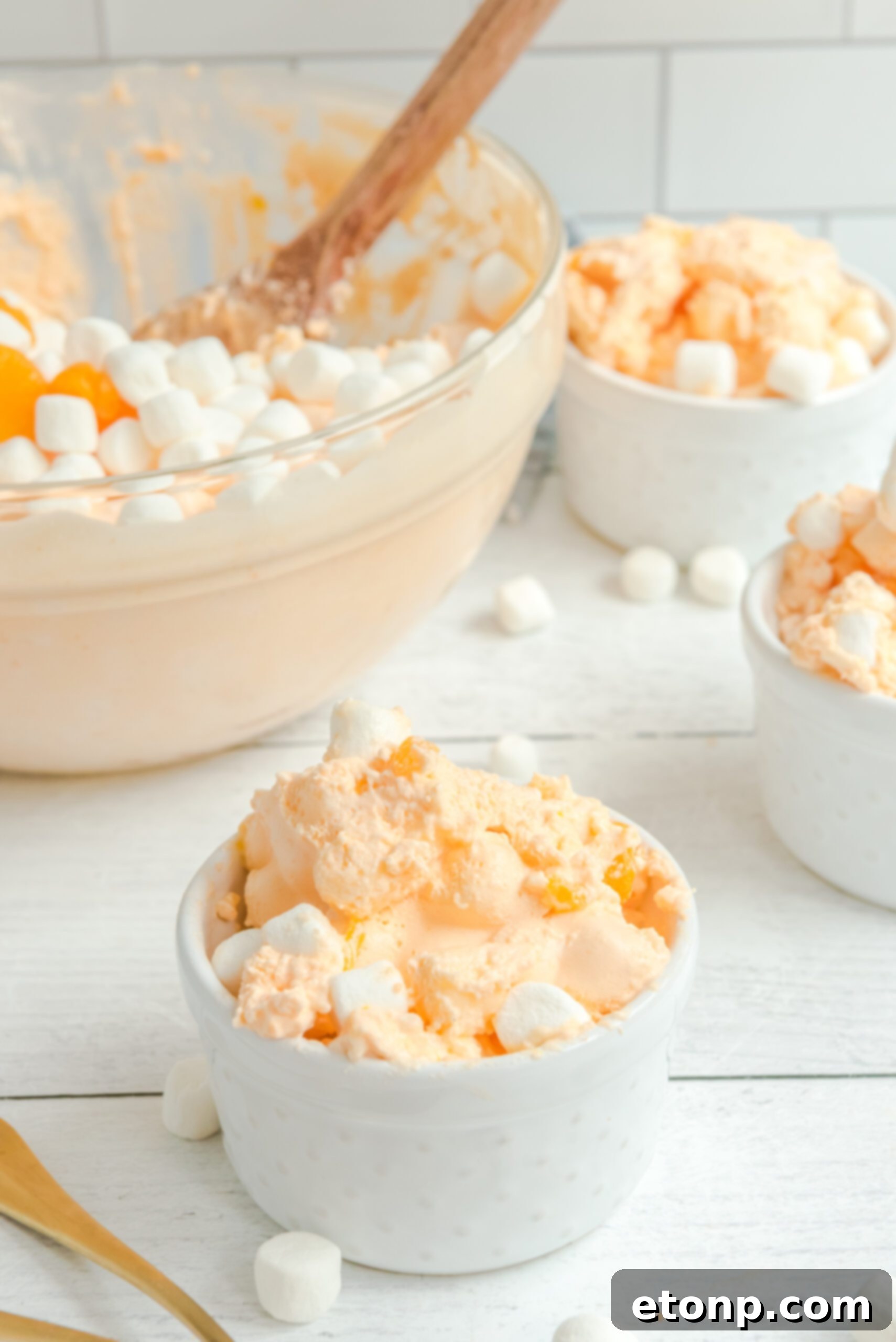 Three small white bowls of Orange Creamsicle Salad, topped with marshmallows and mandarin oranges, with a large glass serving bowl in the background.