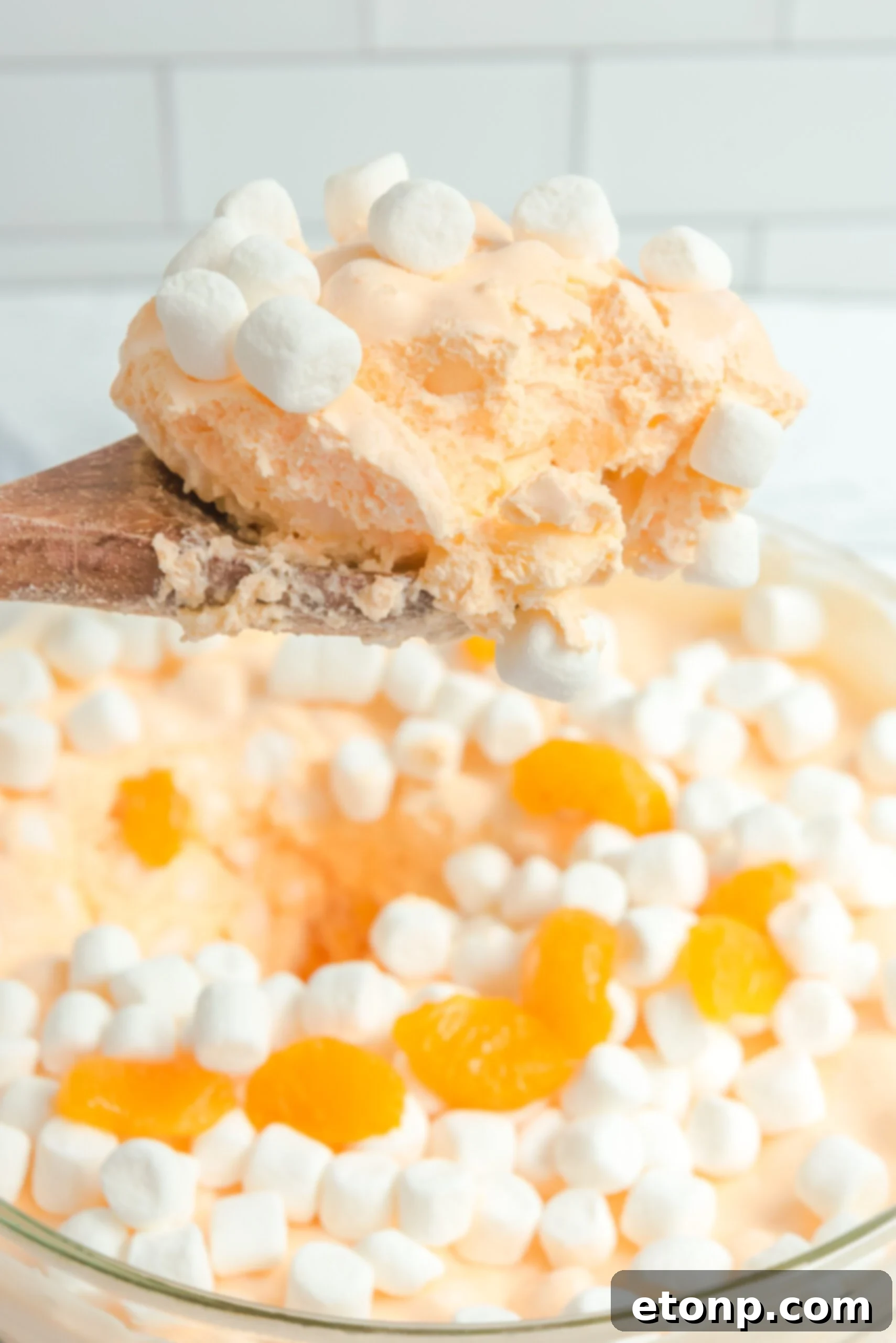 A large serving bowl filled with Orange Creamsicle Salad, with a wooden spoon lifting a scoop, showcasing its fluffy texture.