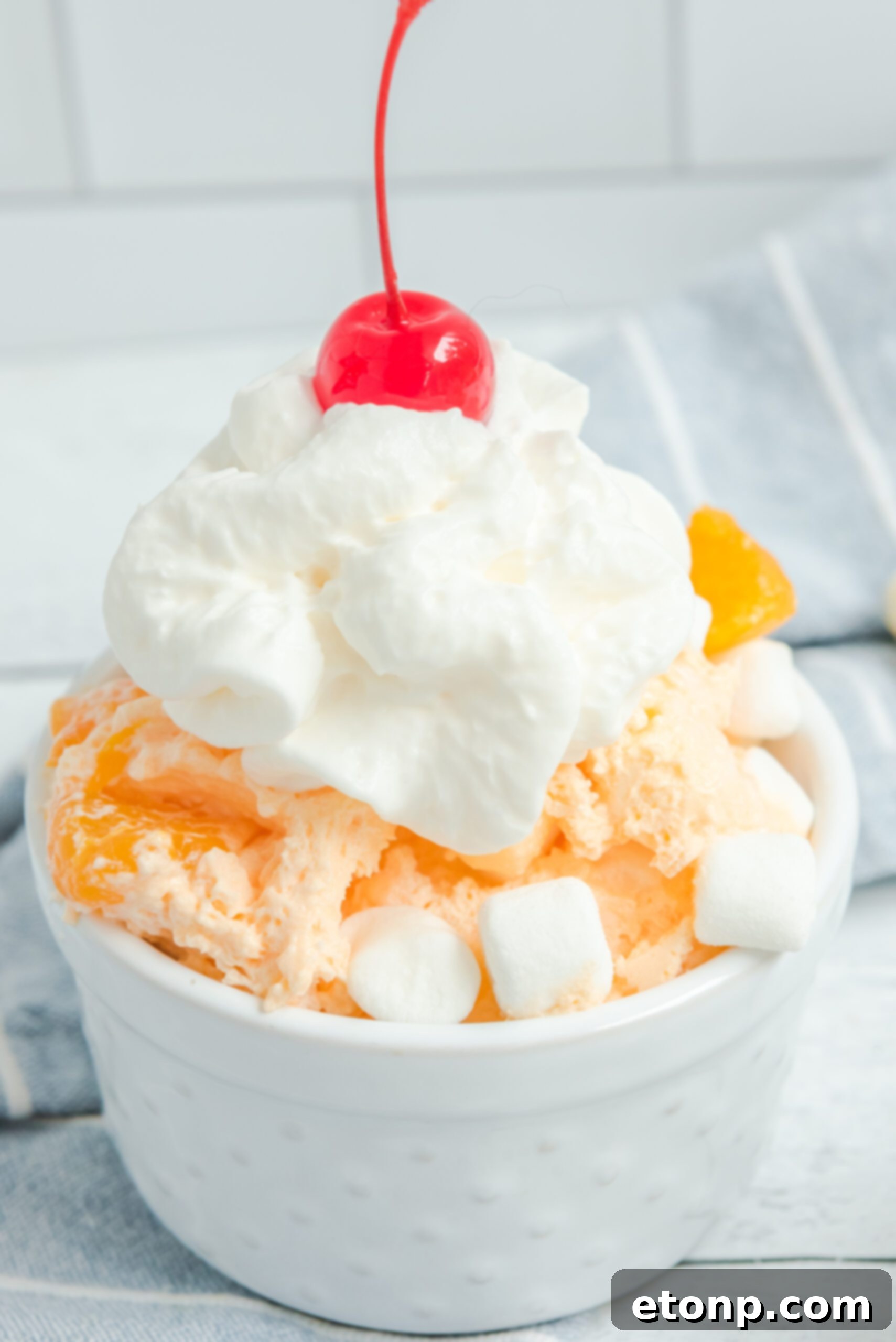 A single serving of Orange Creamsicle Salad in a white bowl, garnished with whipped cream and a bright red cherry.