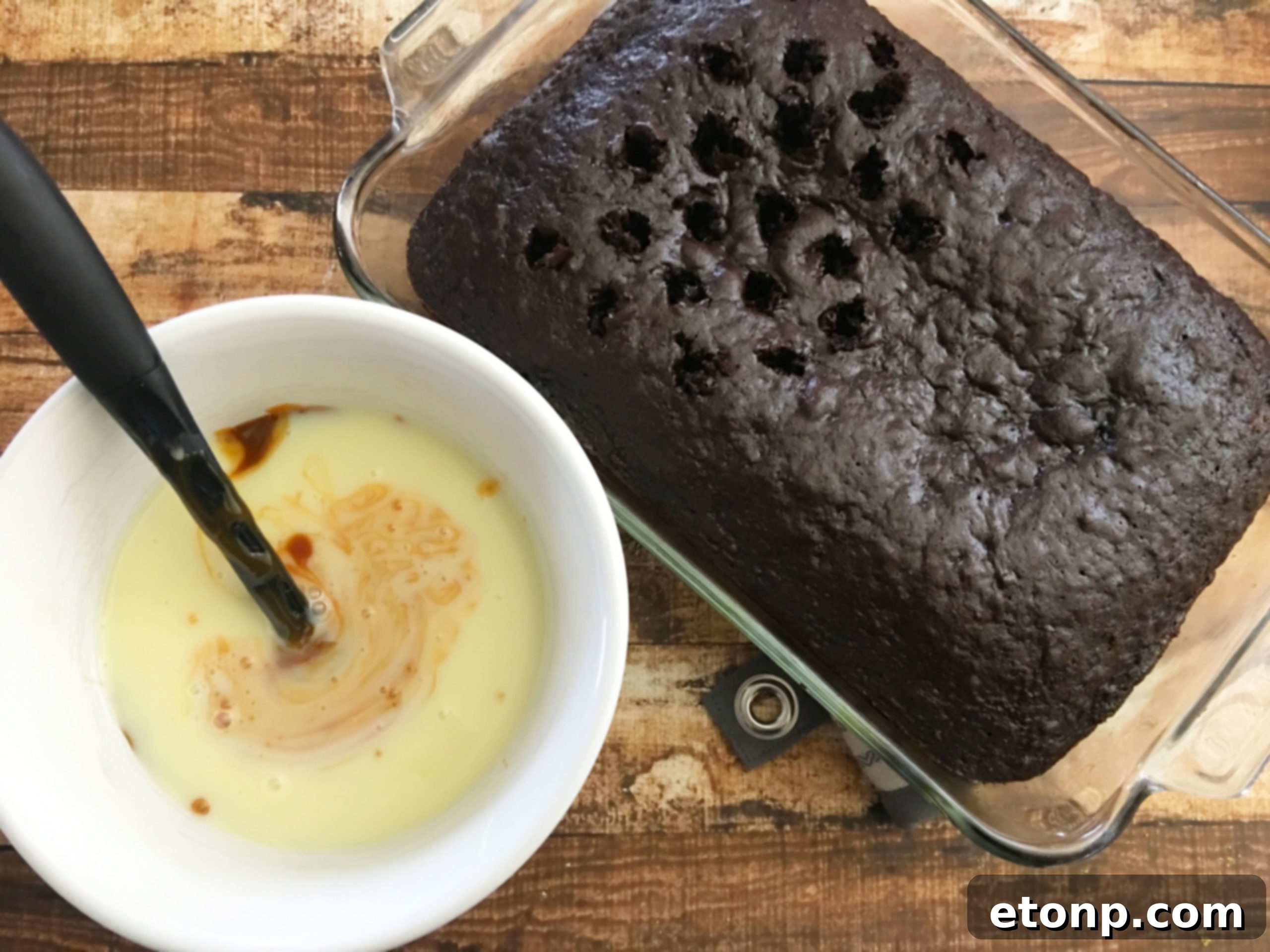 Freshly baked chocolate cake with a wooden spoon handle being used to poke holes into its surface, preparing it for liquid infusion.