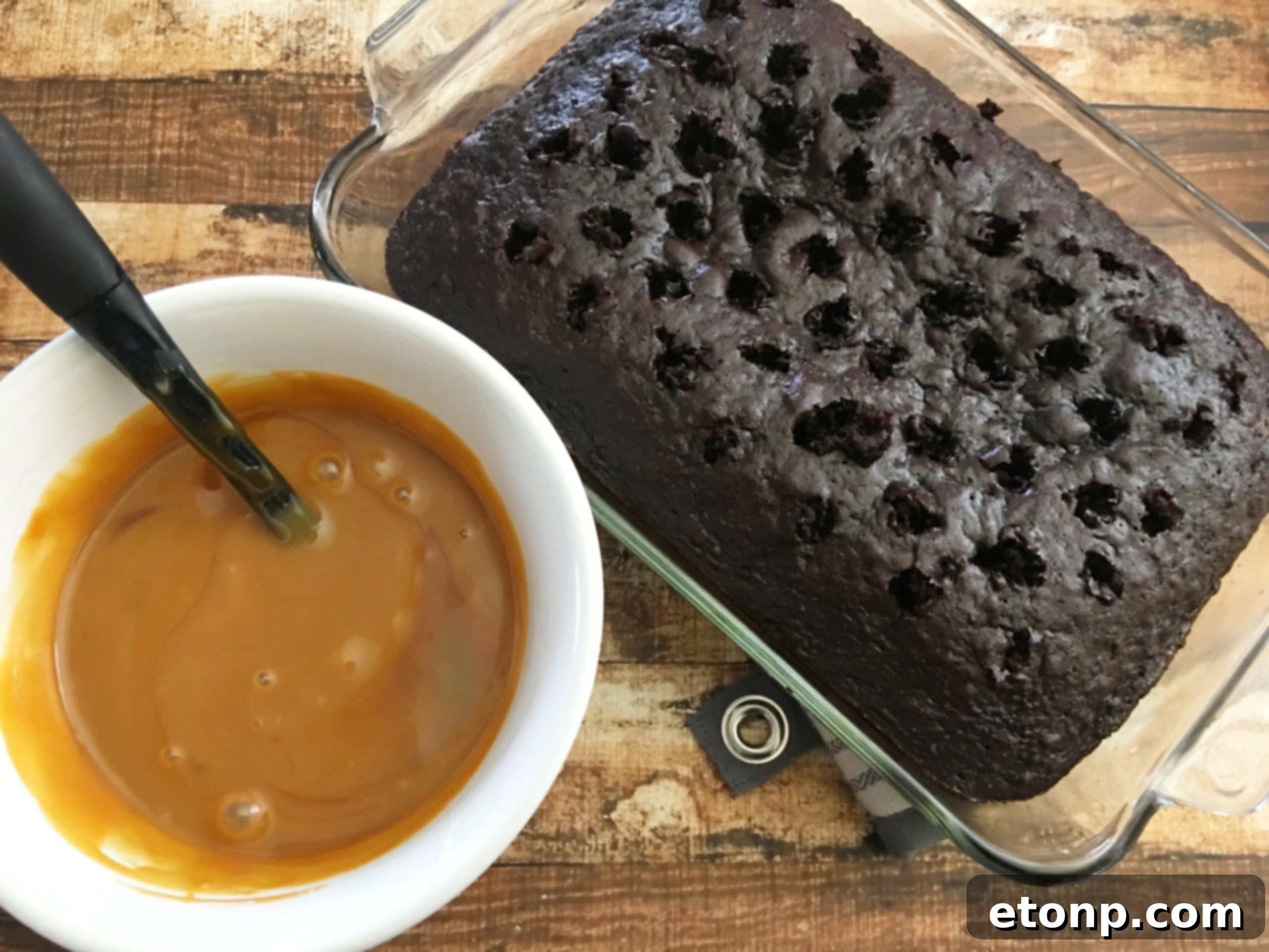 Sweetened condensed milk and caramel mixture being poured slowly and evenly over the poked chocolate cake, allowing it to soak in.