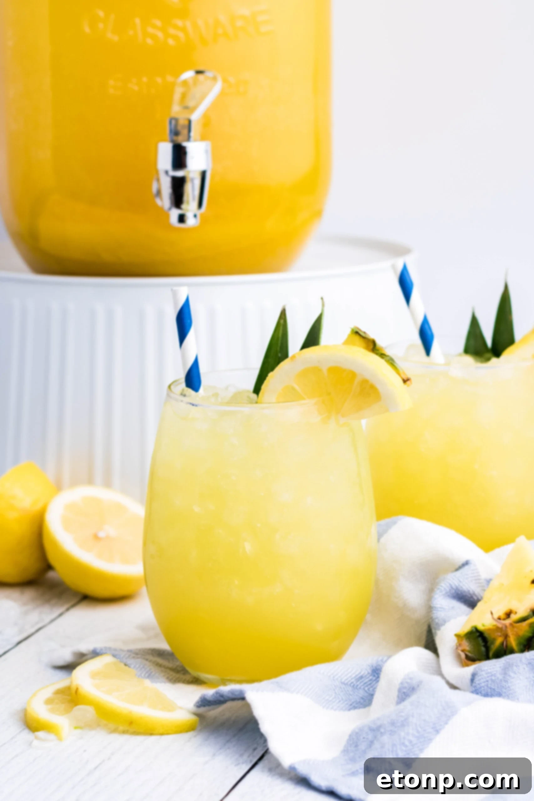Pineapple Lemonade in a clear glass with ice, a blue striped straw, and a fresh lemon slice, shown against a bright background