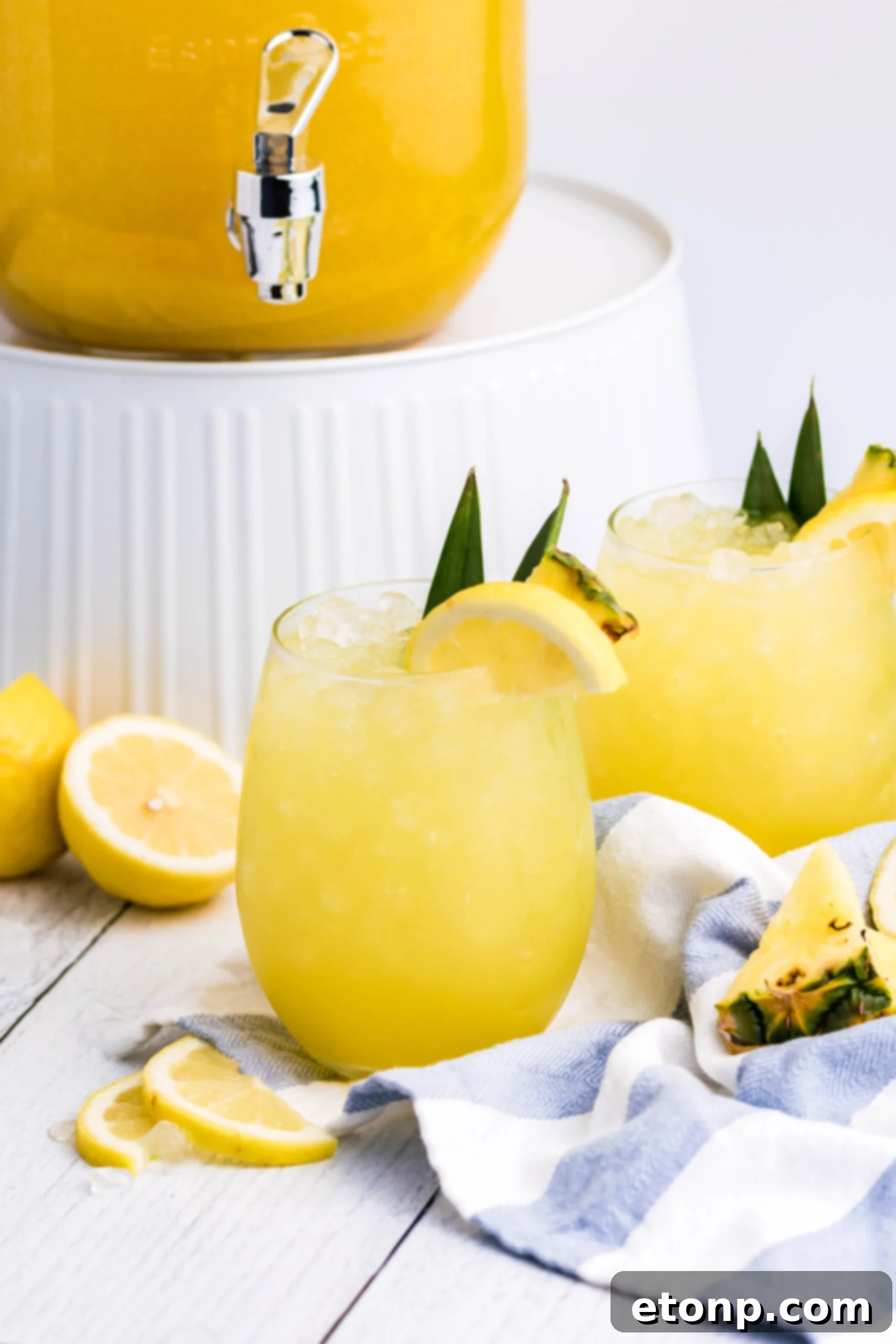 Pineapple Lemonade in a clear glass with ice on a white wood table, with a large punch jar in the soft background