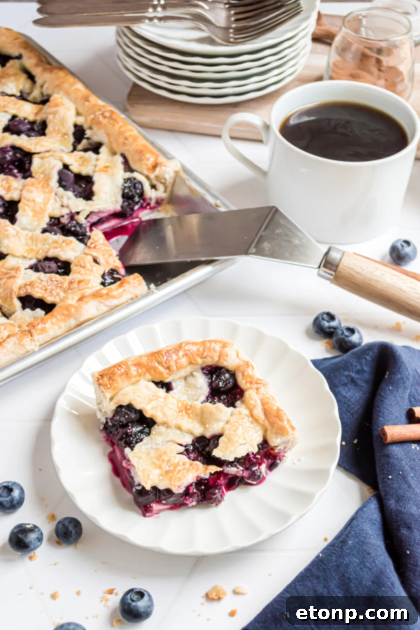 A perfectly baked individual slice of blueberry pie rests on a white plate in the foreground, with the full rectangular slab pie and a silver and wooden spatula visible in the background.