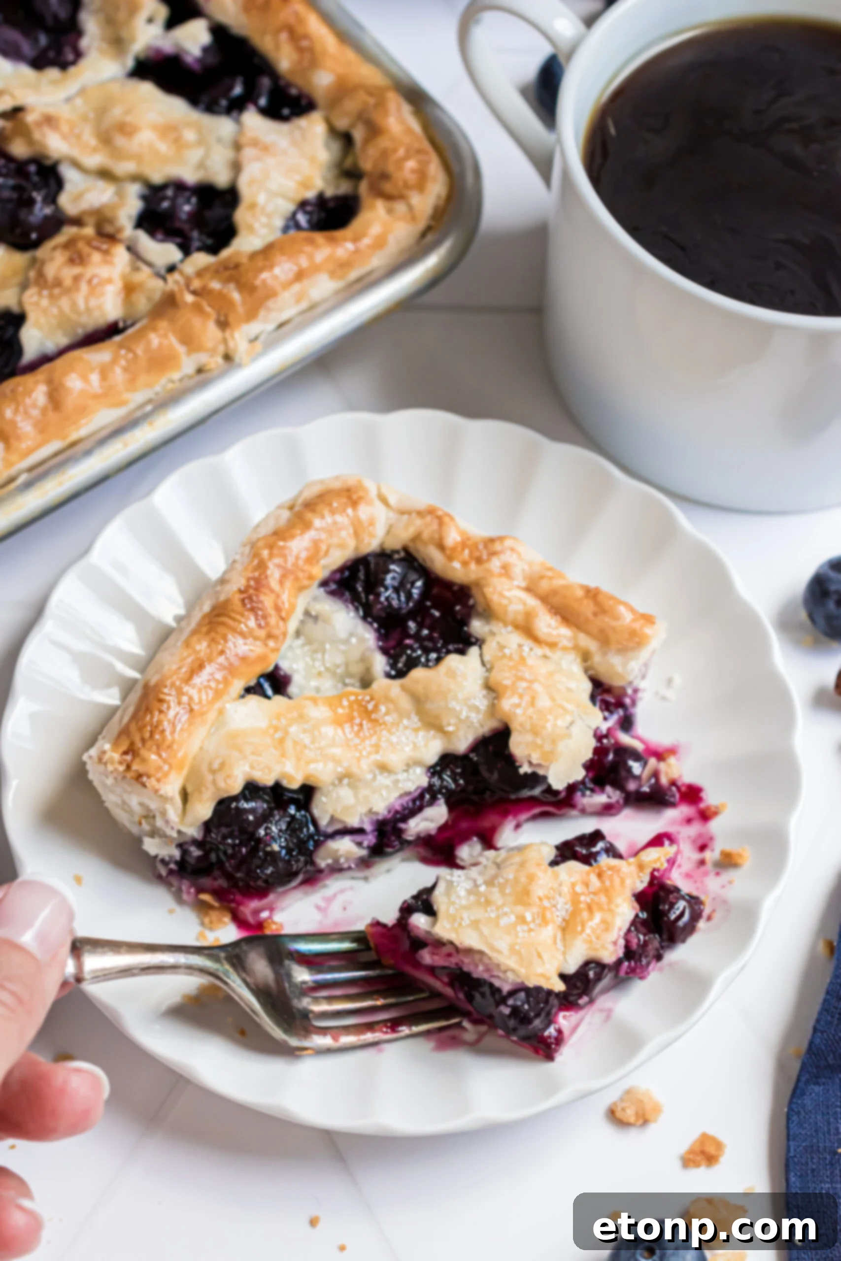 A close-up of a fork gently removing a bite-sized piece of Blueberry Slab Pie from a slice on a white plate, revealing the juicy blueberry filling and golden crust.