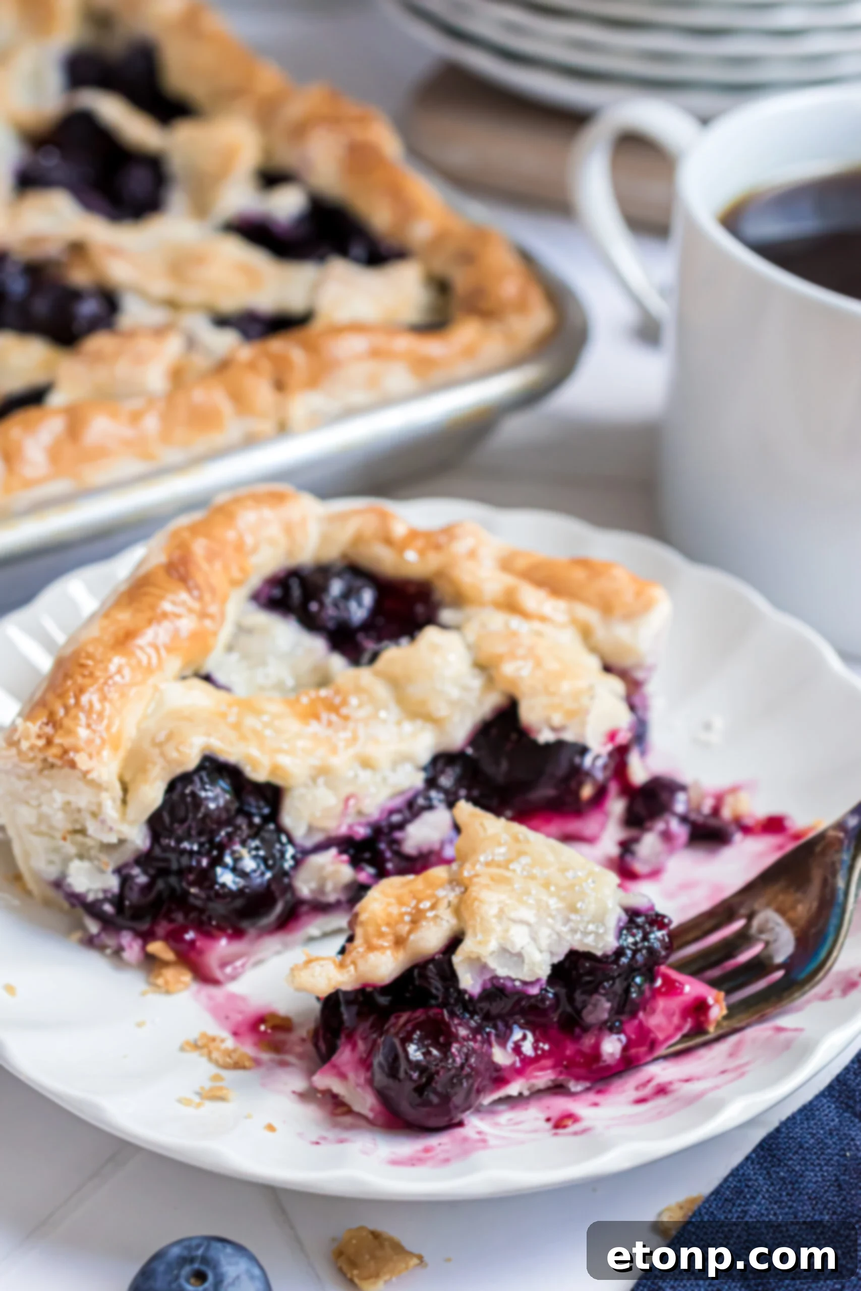 A close-up shot of a single piece of Blueberry Slab Pie on a white plate, with a fork poised to take another delicious bite from the flaky crust and juicy filling.