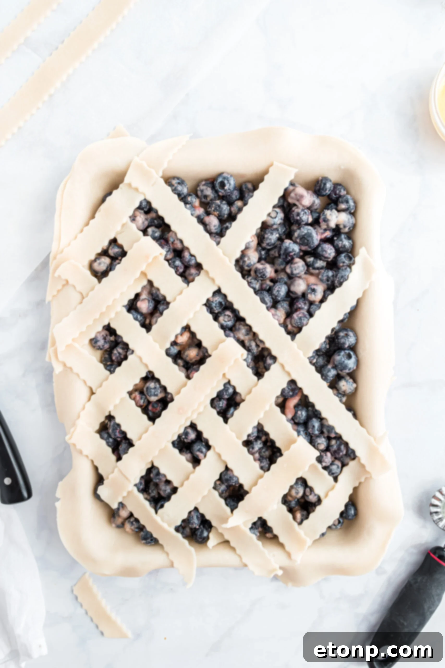A close-up, detailed photograph demonstrating the careful technique for adding the decorative lattice topping onto the blueberry pie, showcasing the woven pattern.