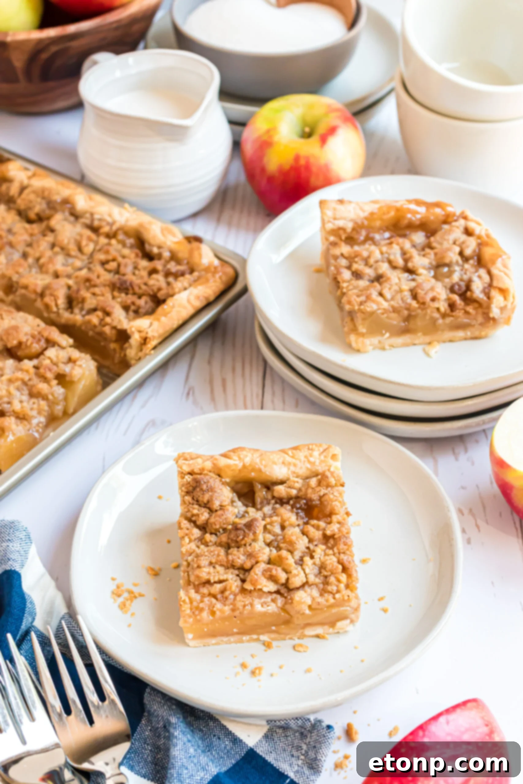 Two slices of apple slab pie on white plates on a white wood background. 