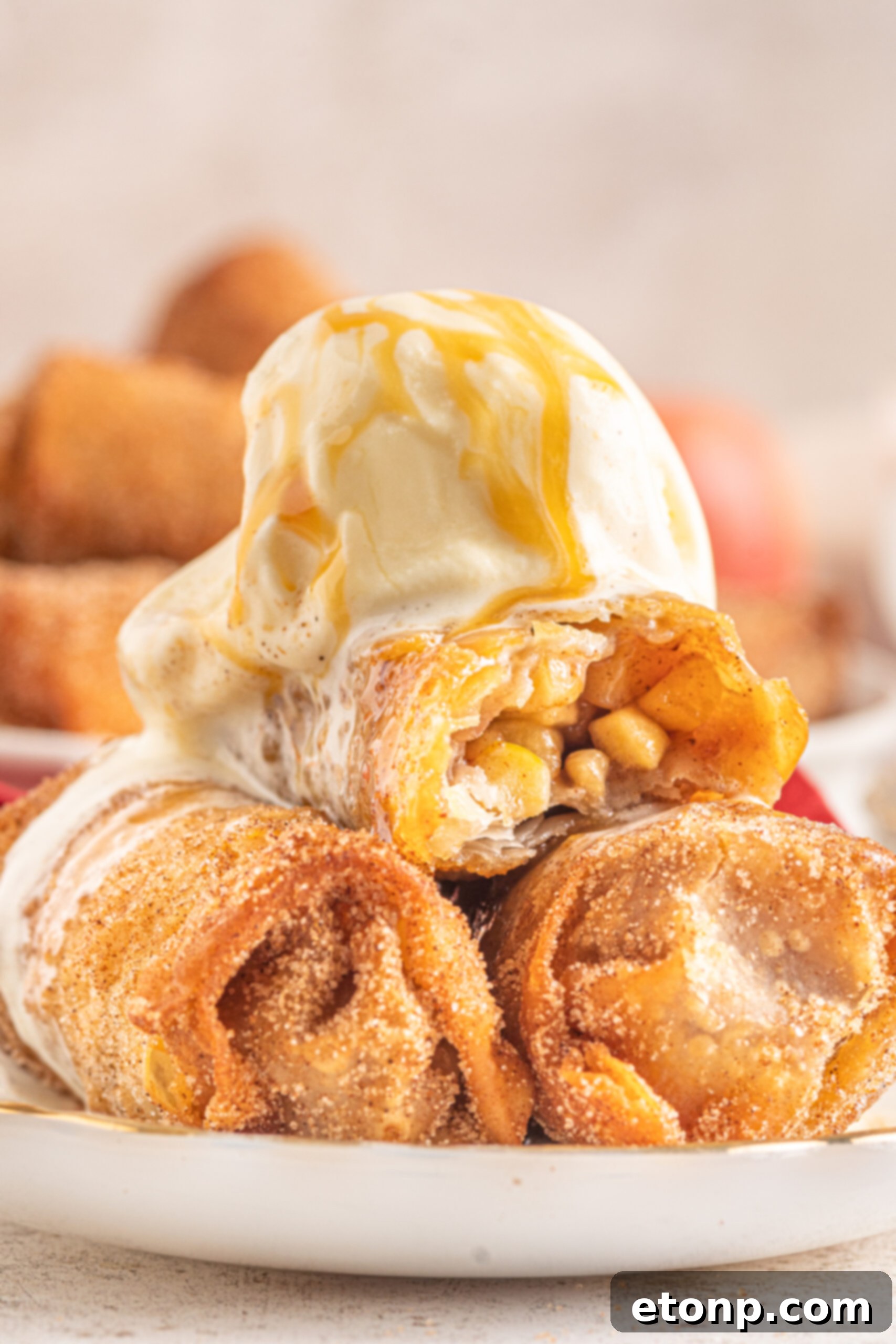 Three golden brown Apple Pie Egg Rolls stacked on a white plate, lavishly topped with melting vanilla ice cream and a rich caramel sauce, ready to be savored.