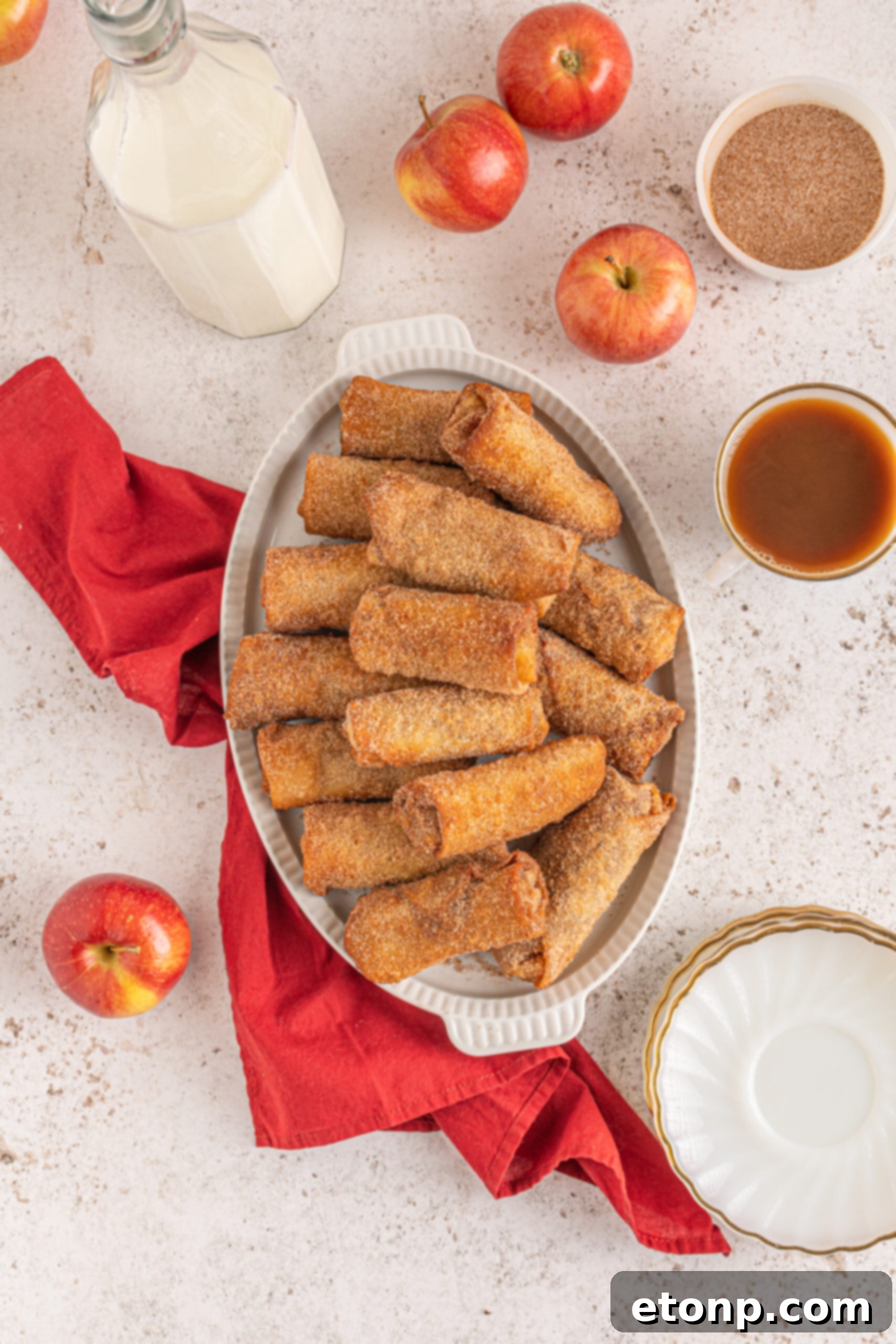A delightful overhead shot showcasing a stack of crispy egg rolls on a clean white platter, with fresh apples and a red linen in the background, creating a charming and mouth-watering presentation.