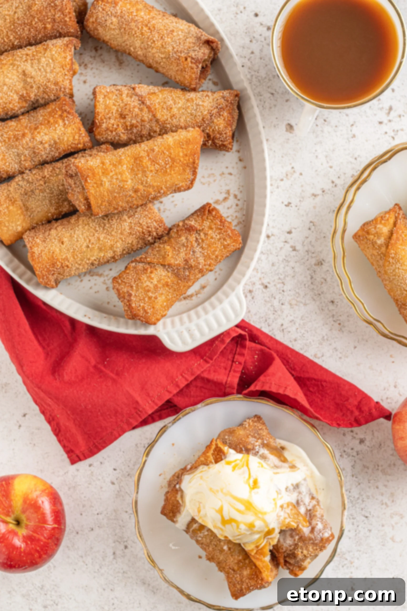 A vibrant overhead shot of delicious Apple pie egg rolls elegantly arranged on a pristine white platter, accompanied by fresh red apples and a contrasting red linen in the background, creating an enticing visual appeal.