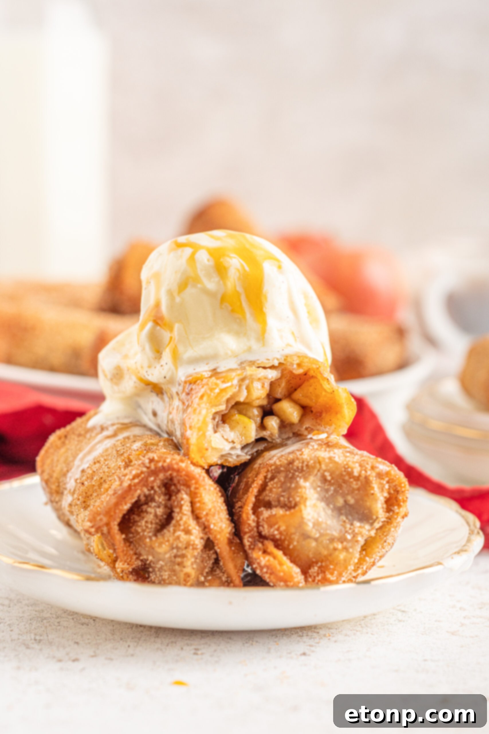 Three perfectly stacked Apple Pie Egg Rolls on a white plate, sumptuously topped with melting vanilla ice cream and a flowing caramel sauce, presenting an inviting and delectable dessert.