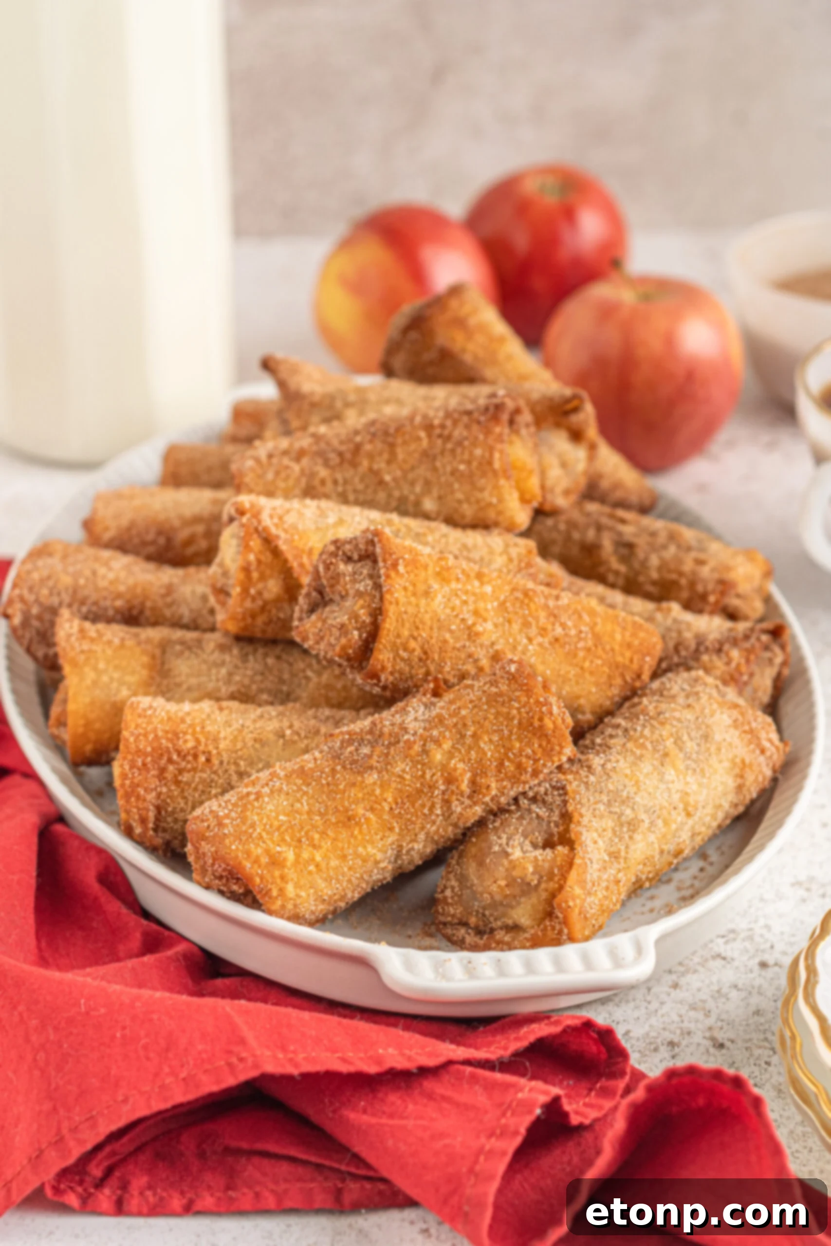 A beautifully arranged platter of Apple Pie Egg Rolls, stacked neatly on a white dish, with fresh red apples and a rustic red linen artfully placed in the soft-focus background, highlighting their delicious appeal.