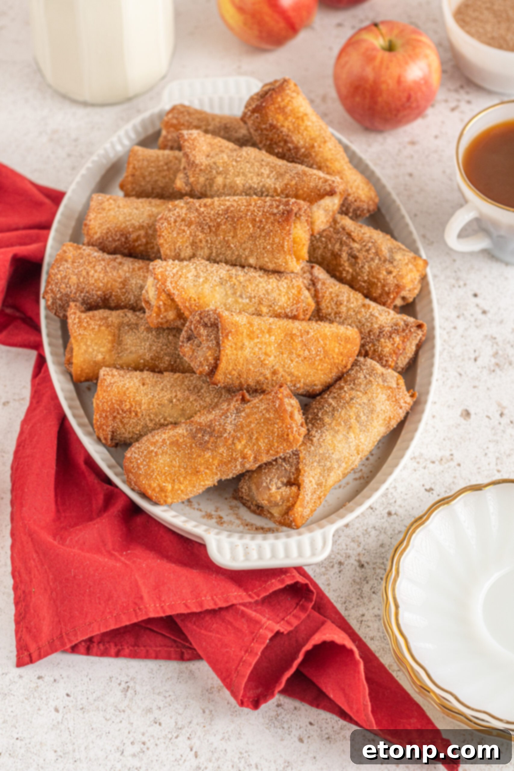 An inviting image of golden-brown egg rolls gracefully stacked on a pristine white platter, artfully arranged with fresh, vibrant red apples and a charming red linen in the background, creating a rustic and appetizing display.