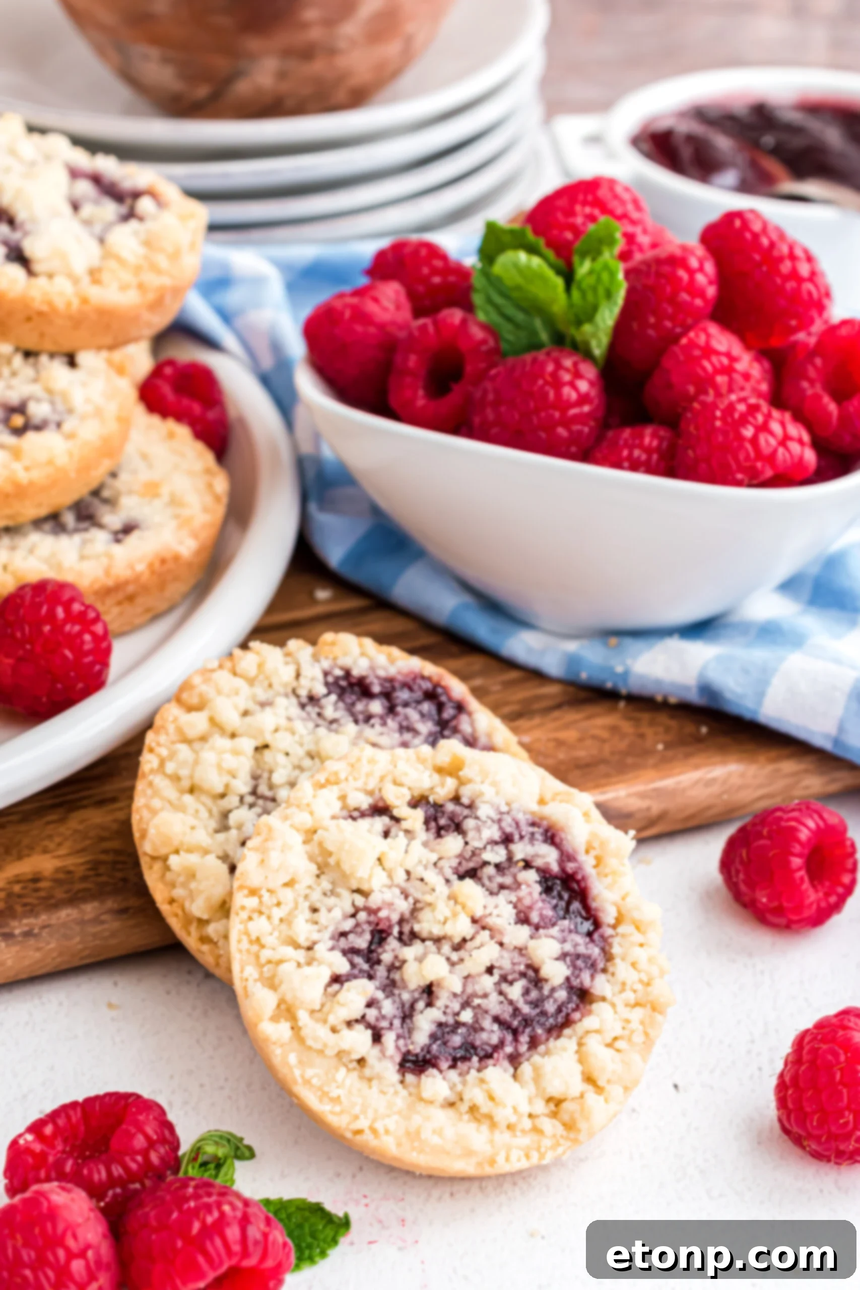 Two raspberry cookies propped against a wooden cutting board with fresh raspberries in a white bowl.