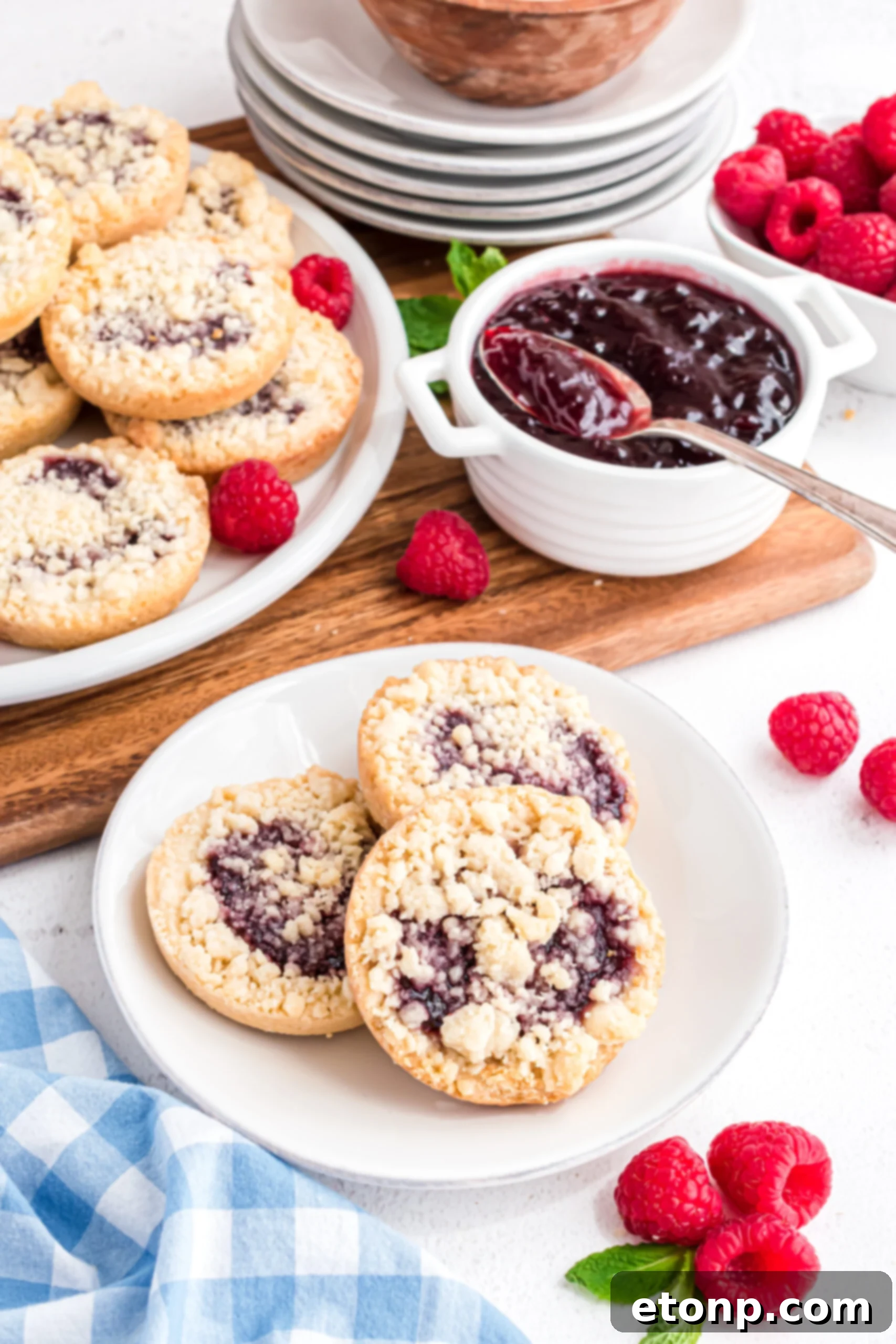 Three raspberry cookies on a white plate with a blue and white linen and fresh raspberries in the background.