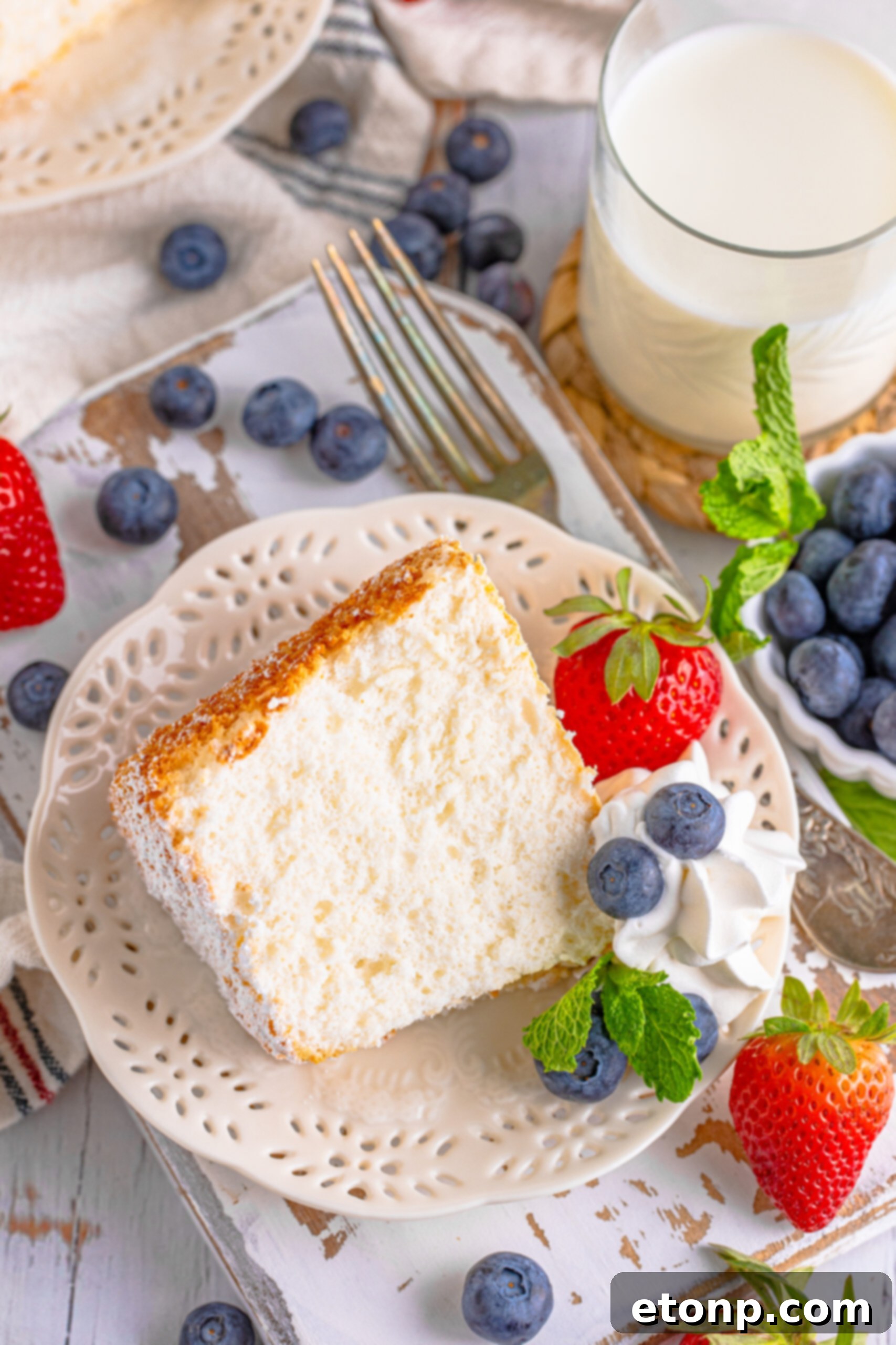 A piece of Angel Food Cake on a white plate with whipped cream, fresh berries, and mint. 