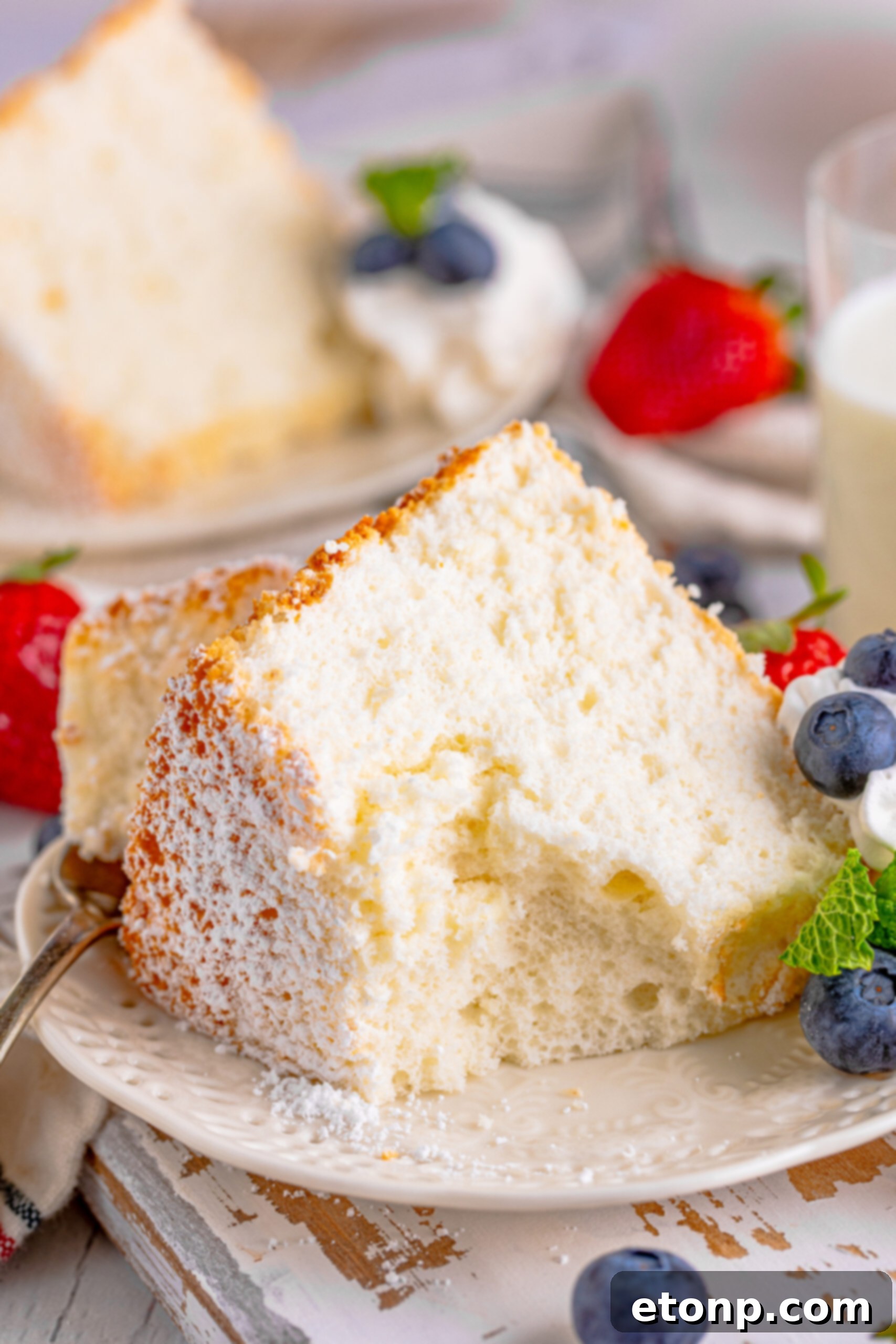 Close up of a slice of Angel Food Cake with a bite taken out of the cake, on a white plate with whipped cream, fresh berries, and mint